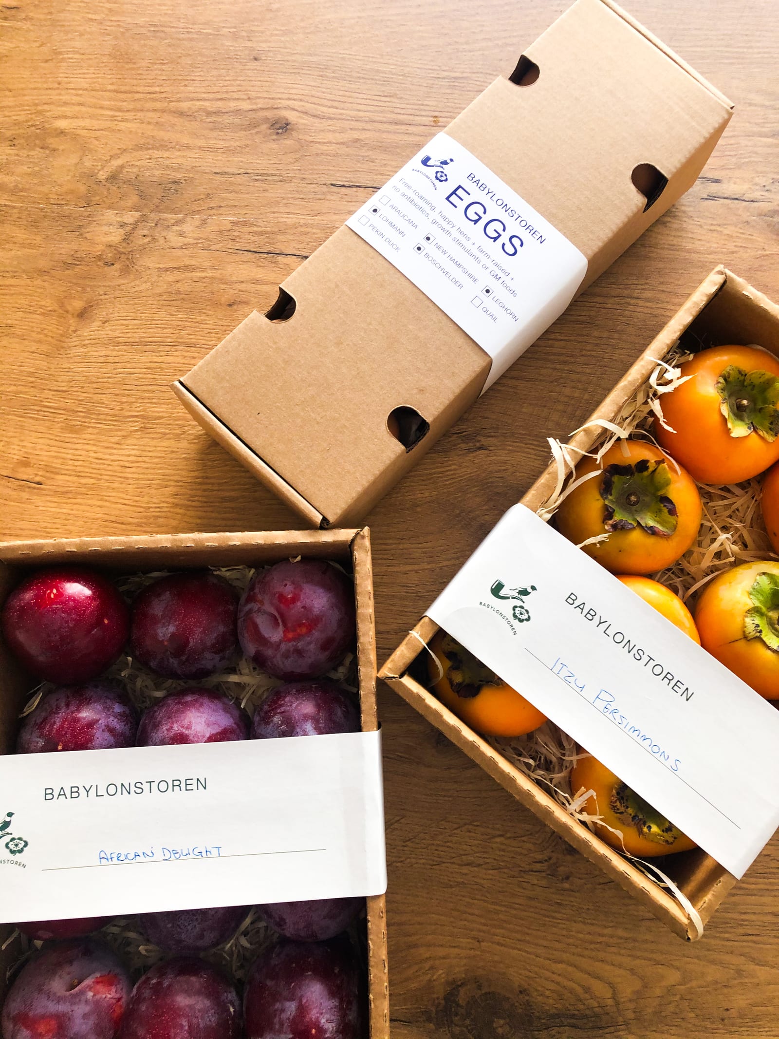 Farm shop produce in Babylonstoren boxes: a carton of eggs, African Delight plums and Fuyu persimmons on a wooden table