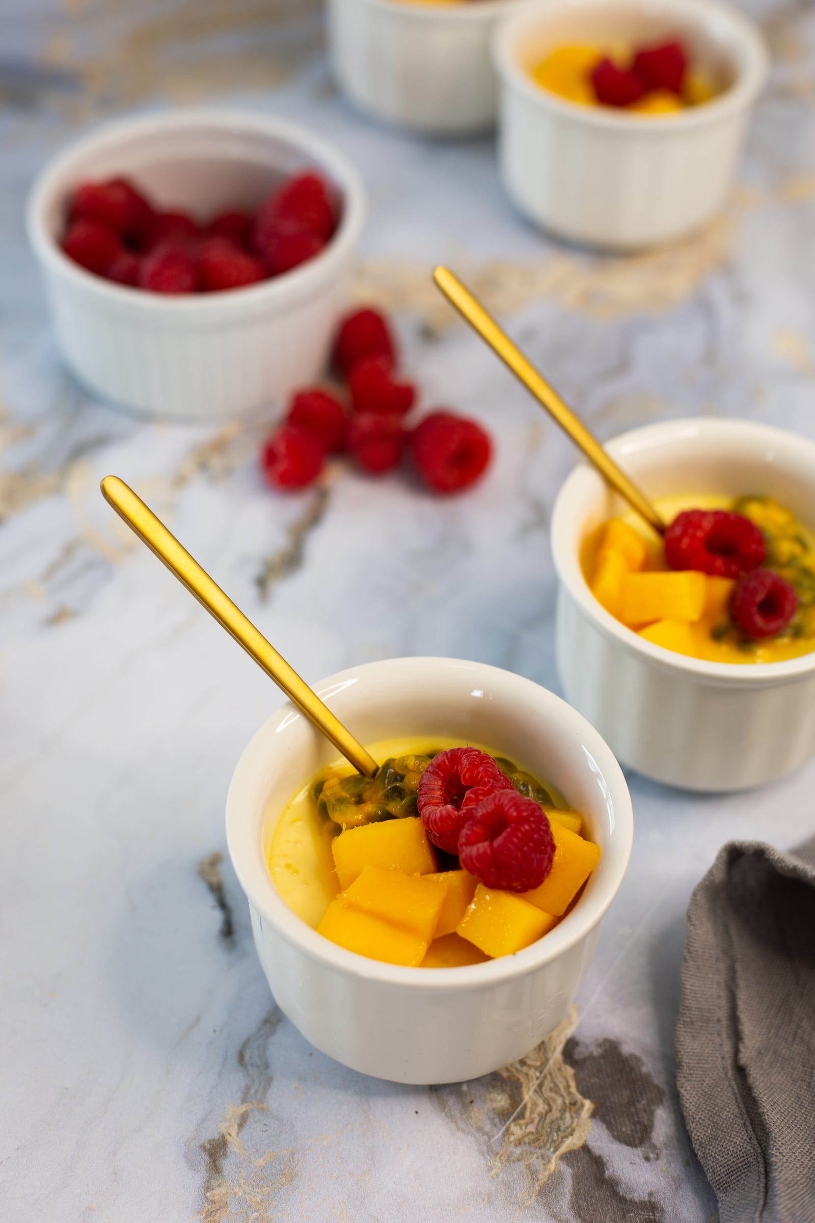 Ramekins of coconut, mango and granadilla baked yogurt on marble at an angle, with scattered raspberries