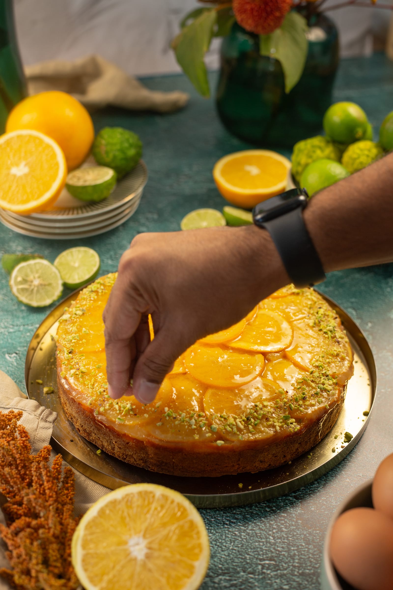 A hand sprinkling chopped pistachios over the orange-topped cake