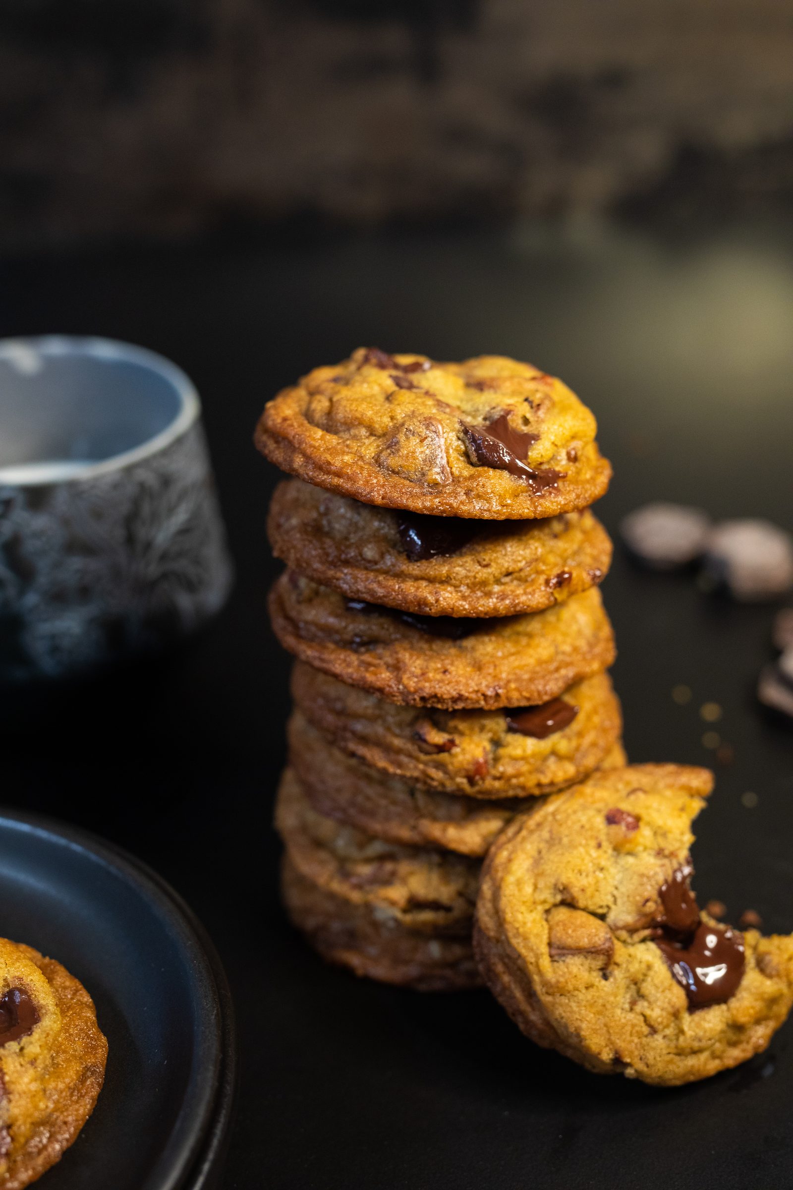 A tall stack of chocolate chip cookies with a broken cookie leaning on it, melted chocolate oozing out, from the side