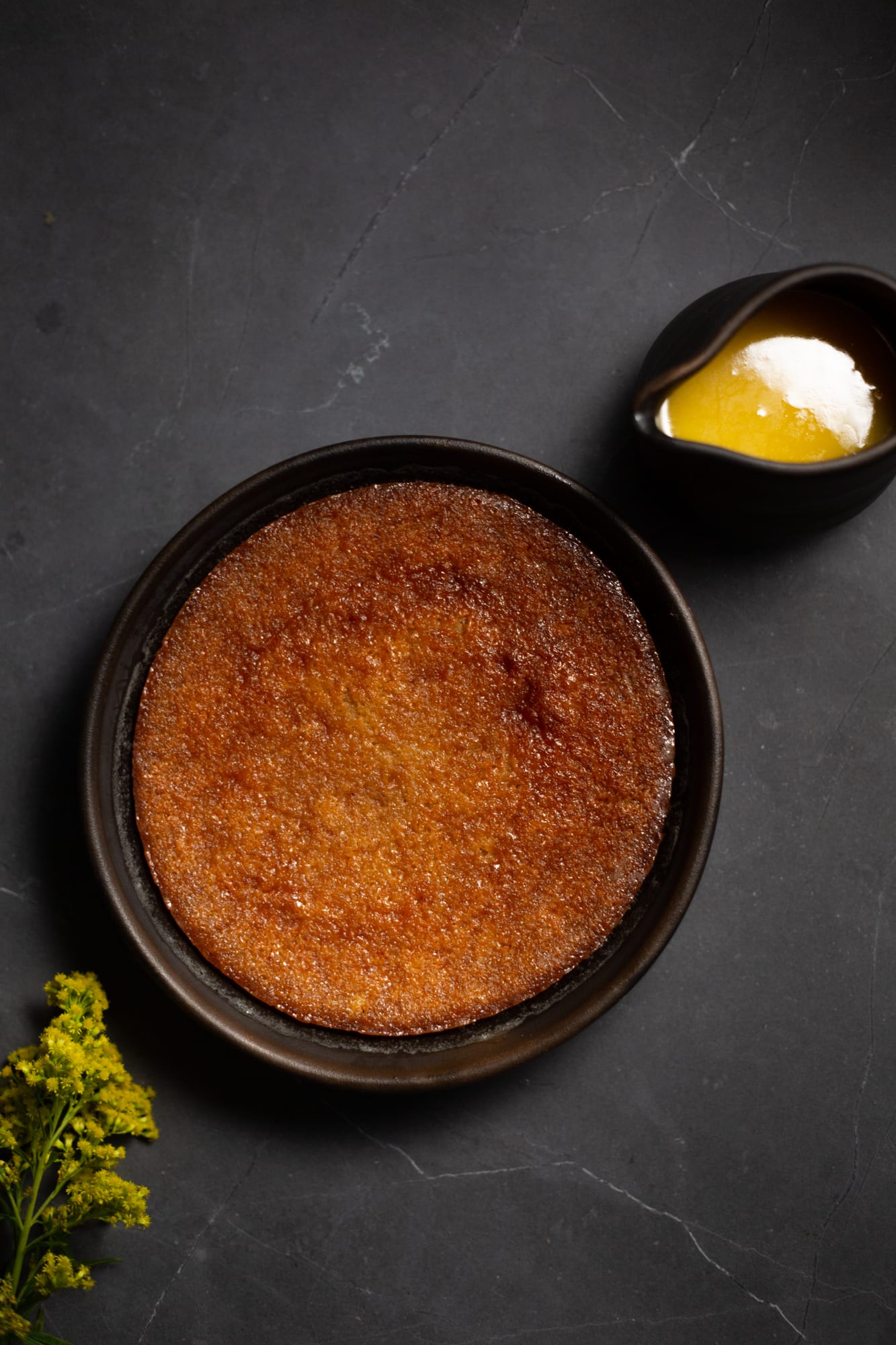 A baked malva pudding in a dark dish, with a jug of crème anglaise, from above