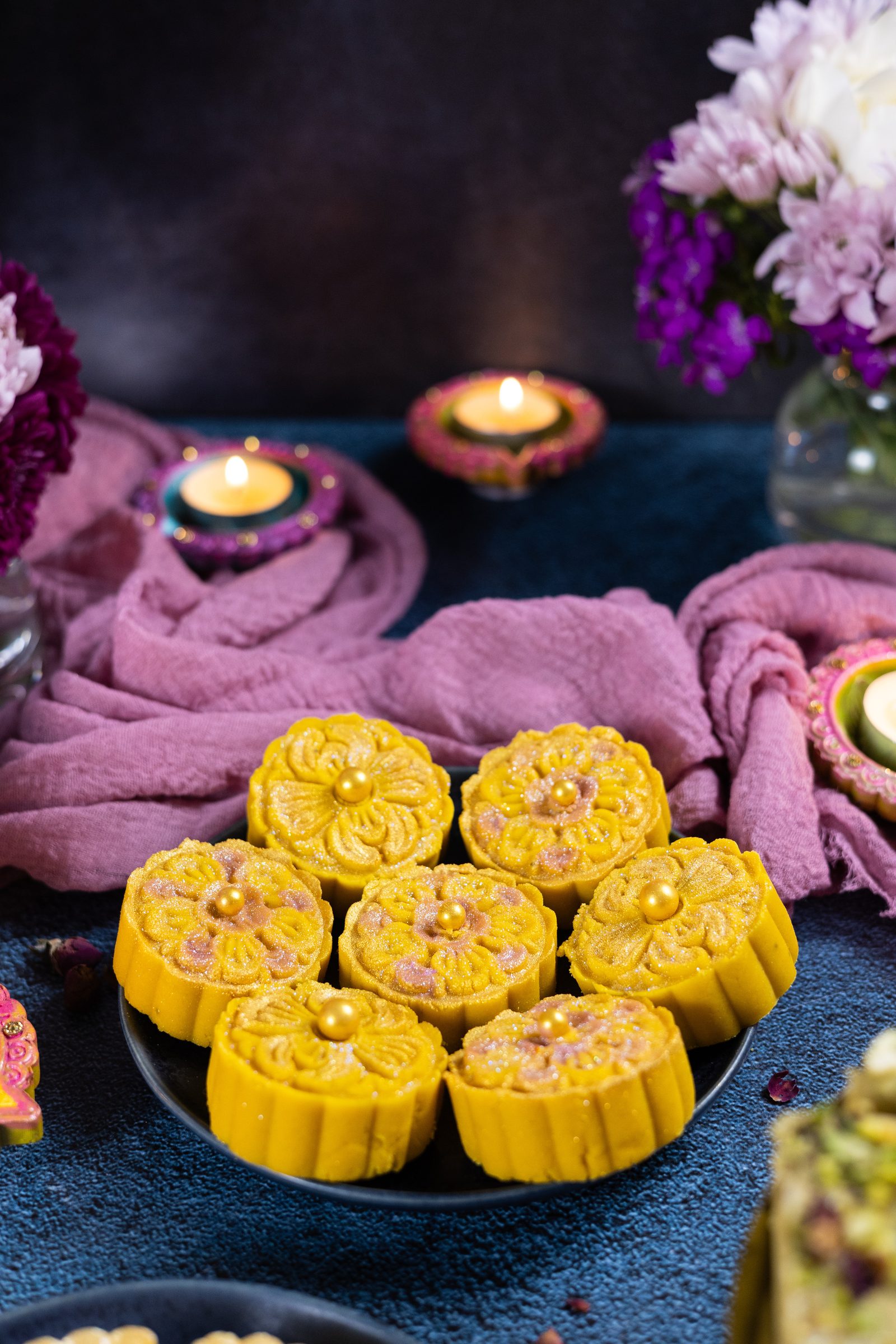 Flower-shaped mango burfee on a dark plate from the side, with candles and flowers behind