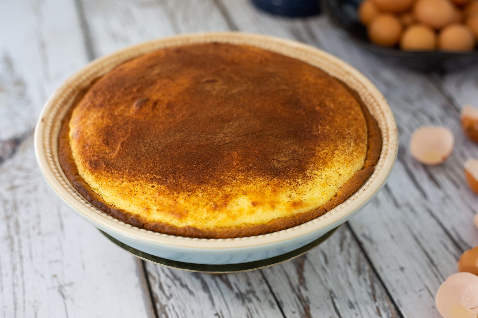 A whole melktert dusted with cinnamon beside eggs and eggshells on white wood