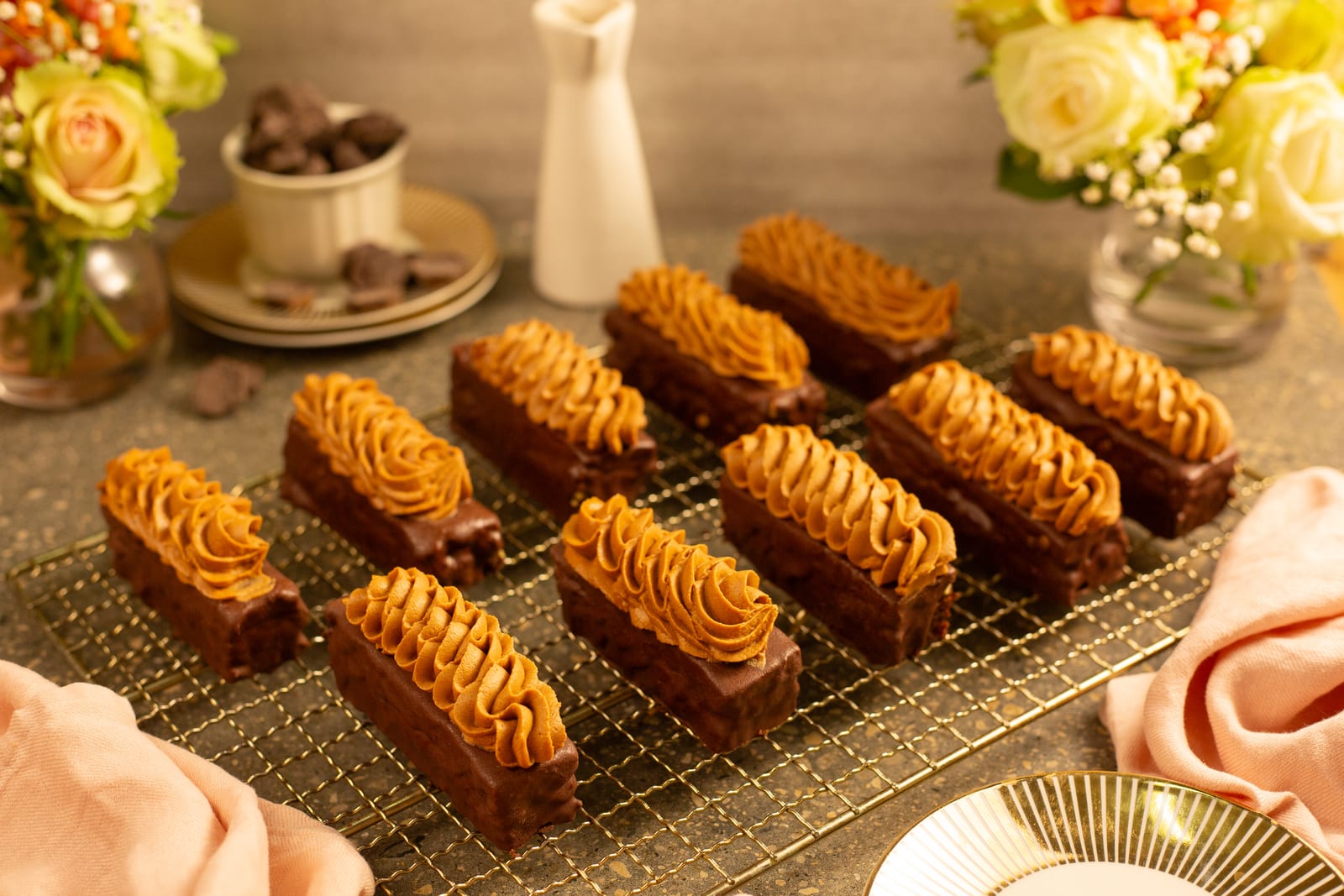Rows of channa magaj and chocolate brownie fingers on a gold wire rack, with flowers and a cup of chocolates