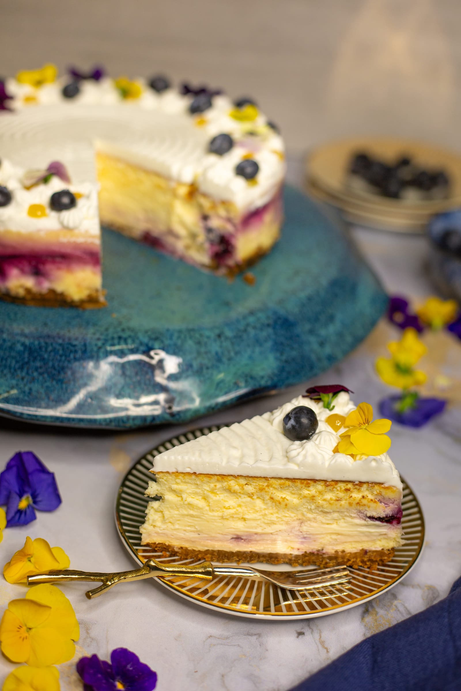 A slice of lemon and blueberry cheesecake on a gold plate in front of the cut cake