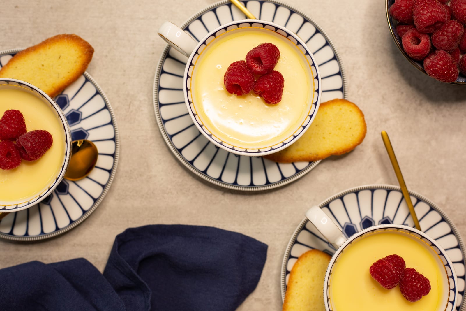 Lemon possets in patterned teacups from above, with raspberries, cat's tongue biscuits and a navy cloth