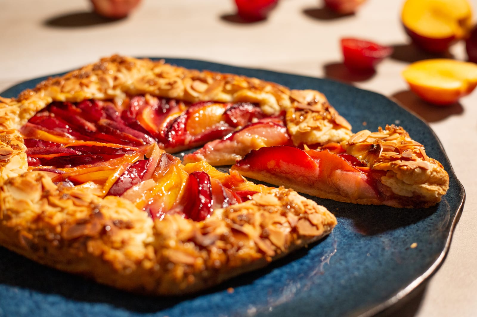 A stone fruit galette with a wedge cut on a teal plate, fruit behind, from a low angle