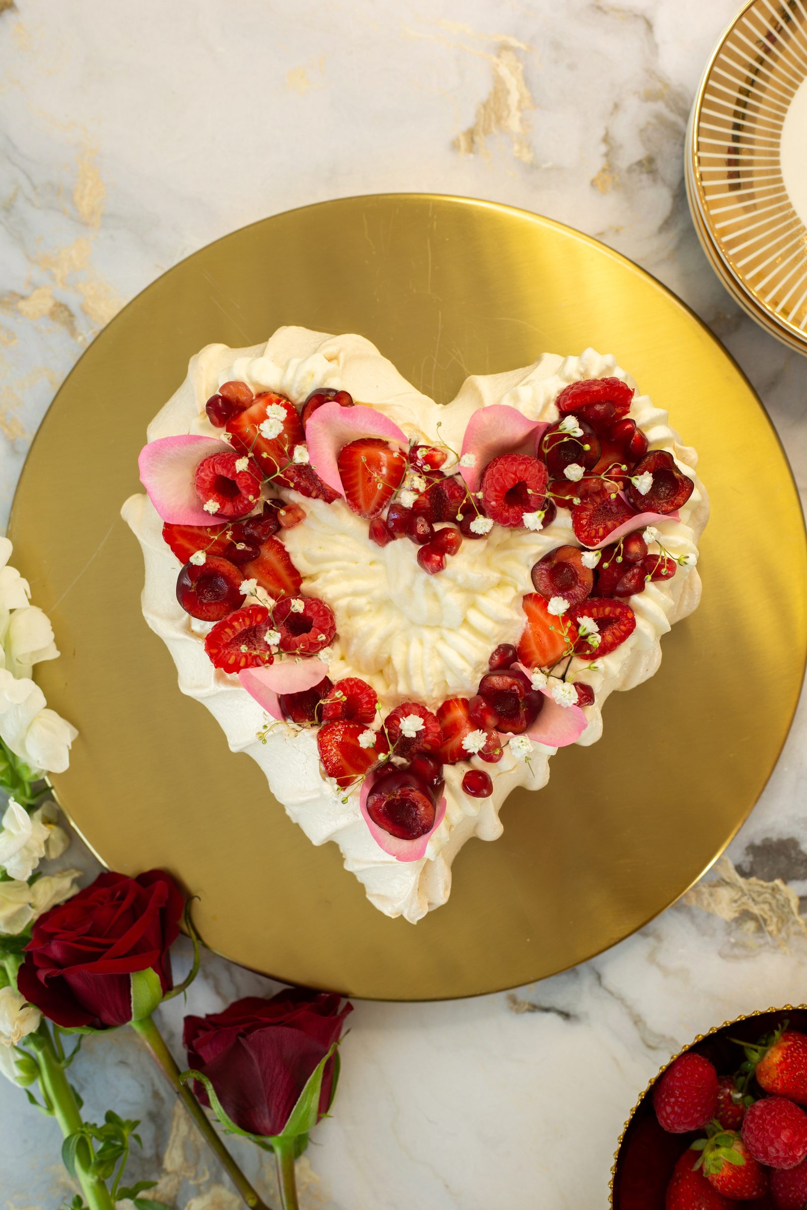 A heart-shaped berry pavlova on a gold board beside red roses and a bowl of strawberries, from above