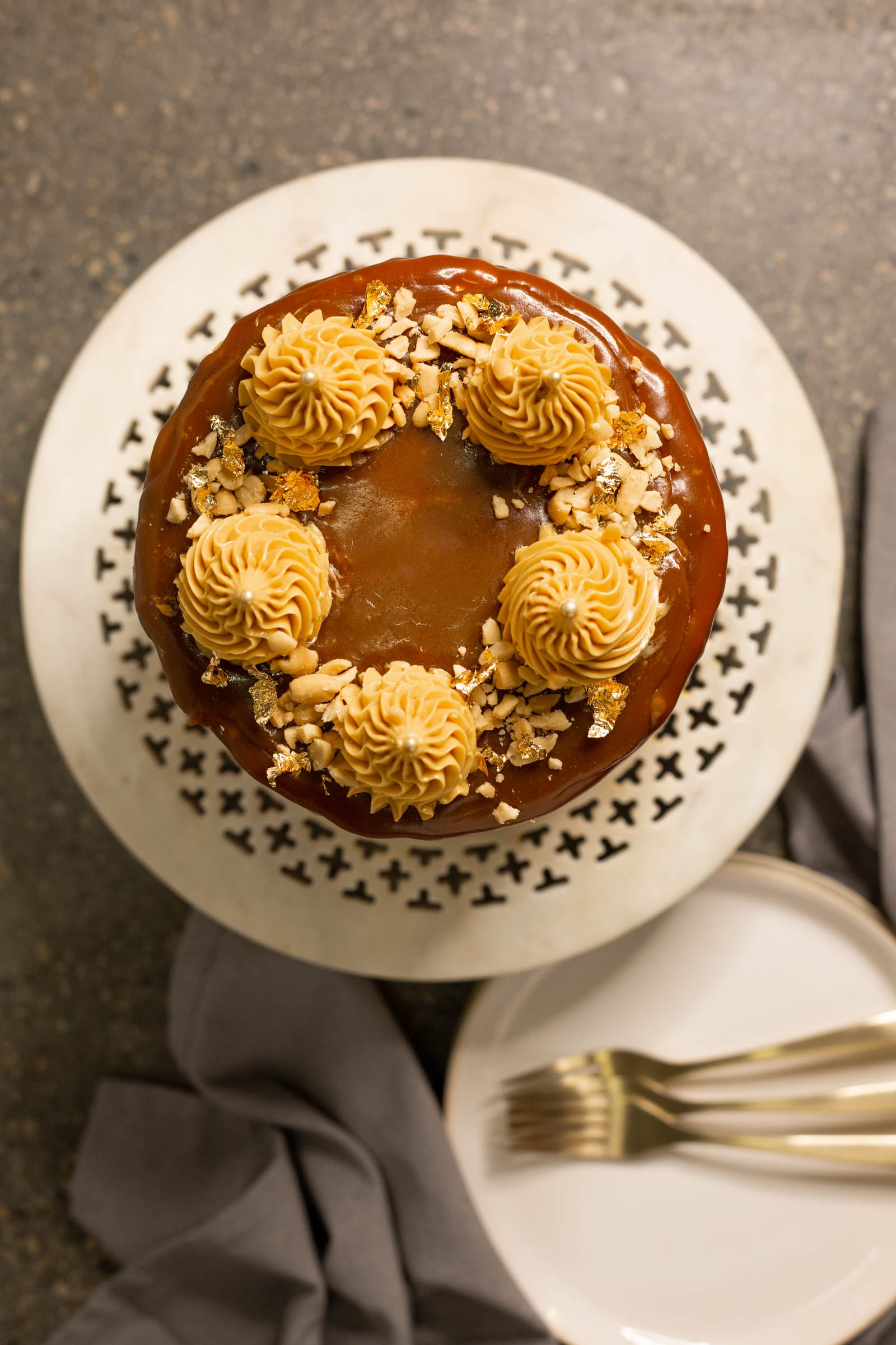 A salted caramel peanut cake on its stand with plates and forks below, from above