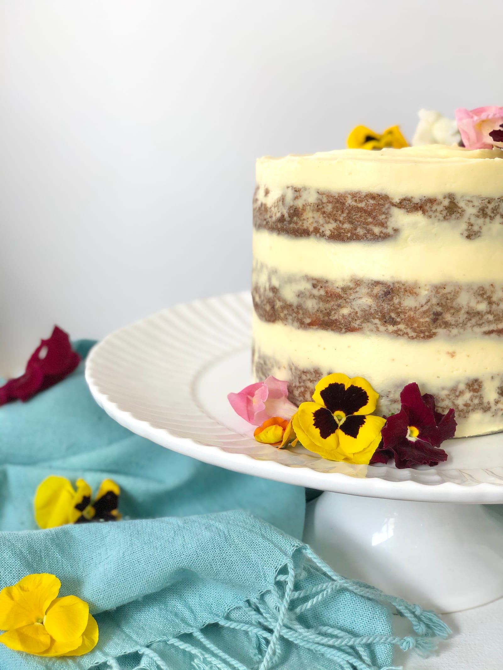 A whole carrot cake on a white cake stand with a teal cloth