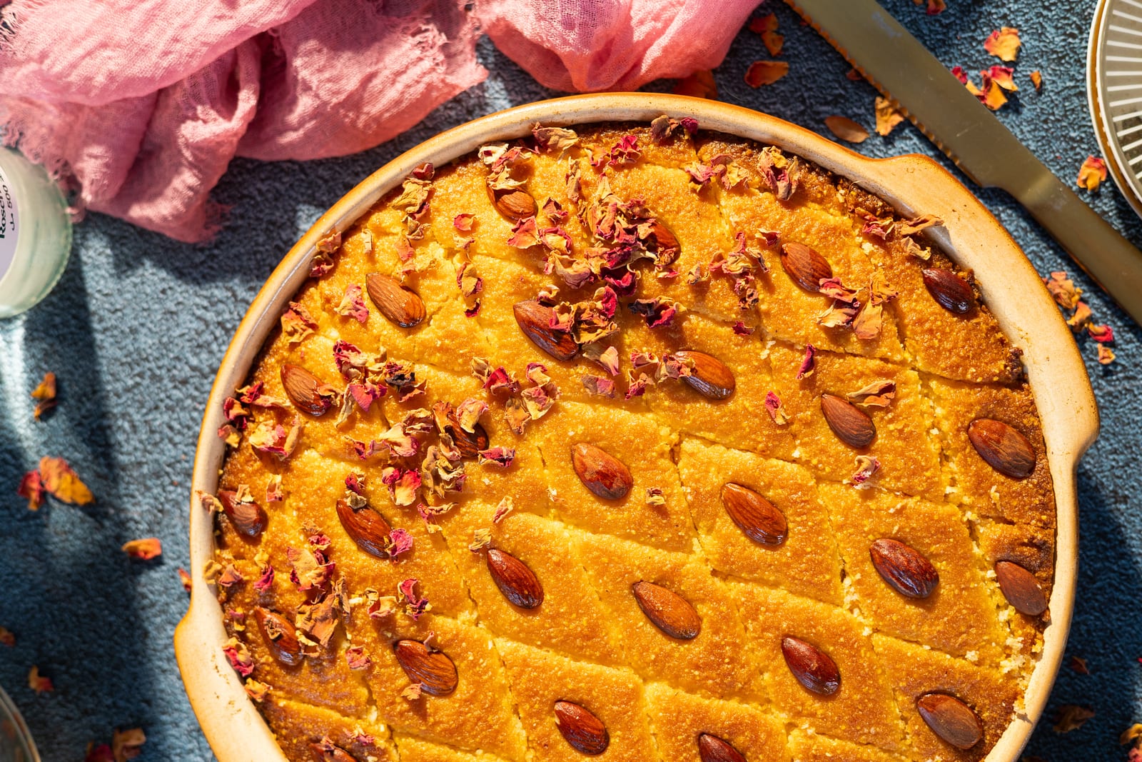 A semolina, coconut and polenta basbousa in a round dish, scattered with rose petals