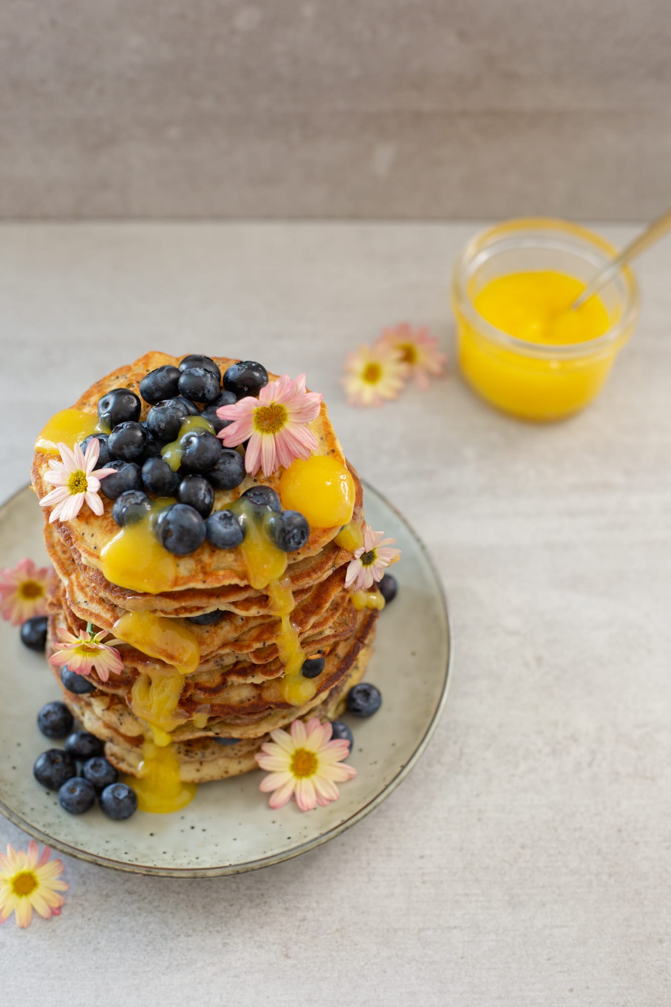 A stack of lemon and poppyseed pancakes topped with lemon curd, jars of curd behind, from above at an angle