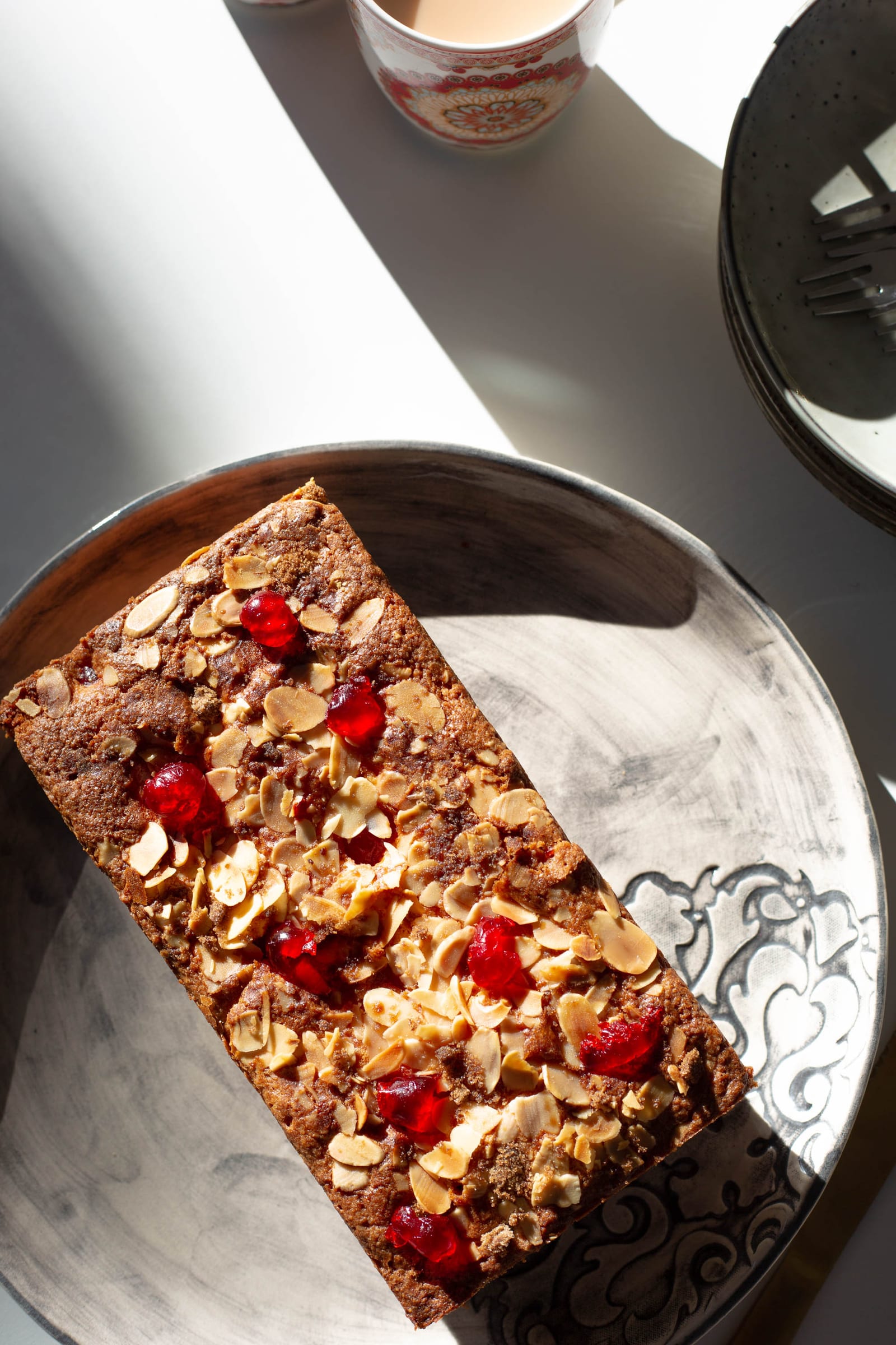 A gluten-free cherry and almond loaf on its plate from above, in strong diagonal shadow