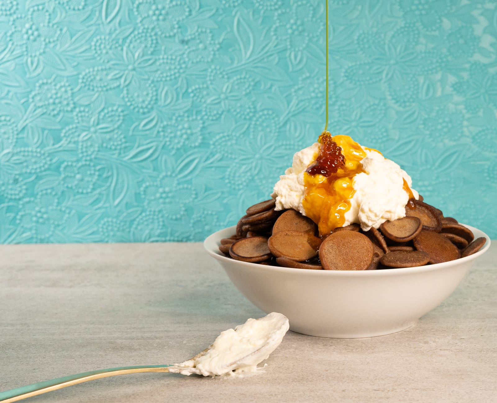 Syrup drizzled over cream on a bowl of Milo pancake cereal against a turquoise wall