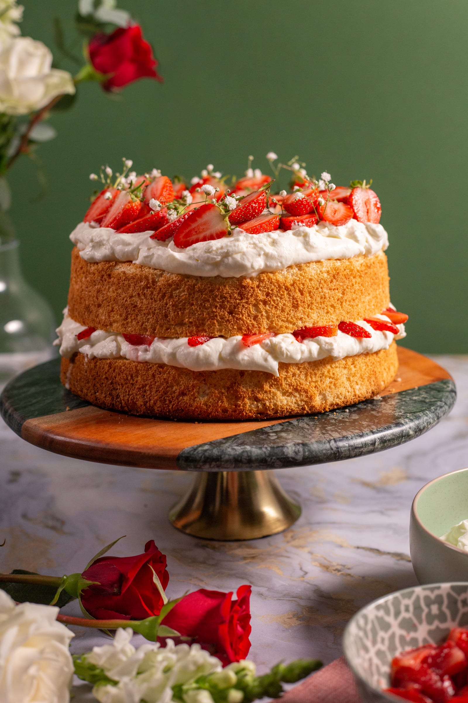 A strawberry and cream angel food cake on a tall gold cake stand against a green wall, roses beside it