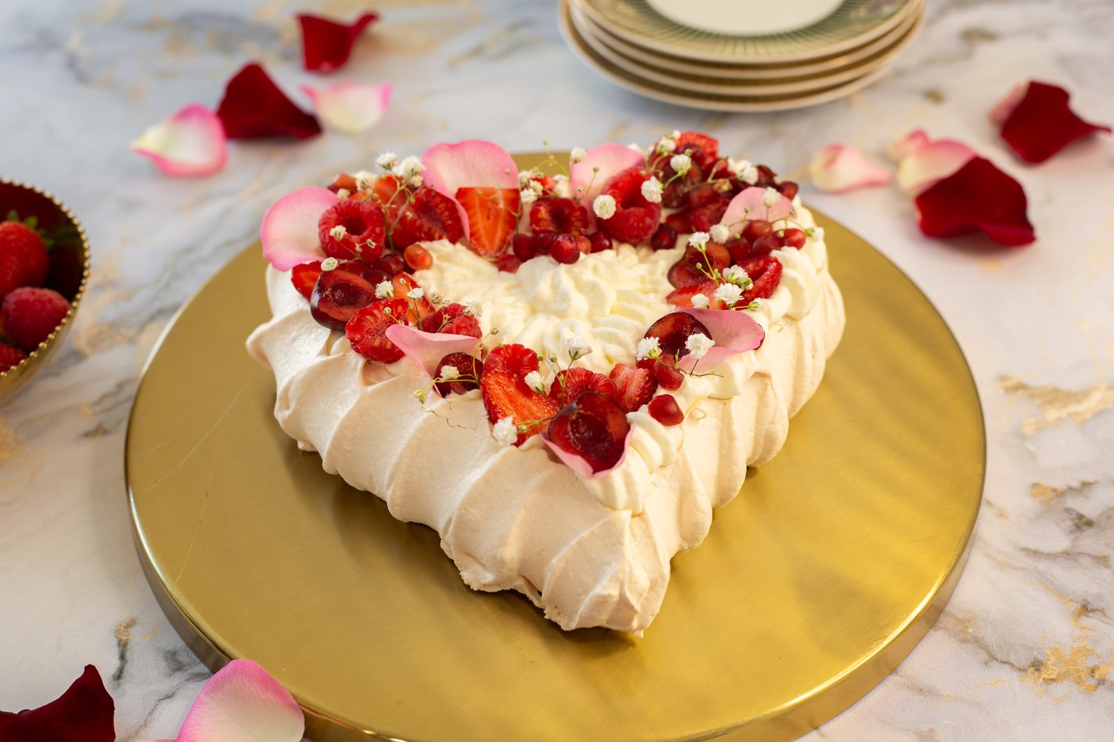 A heart-shaped berry pavlova showing its meringue sides, with plates and rose petals, from a low angle