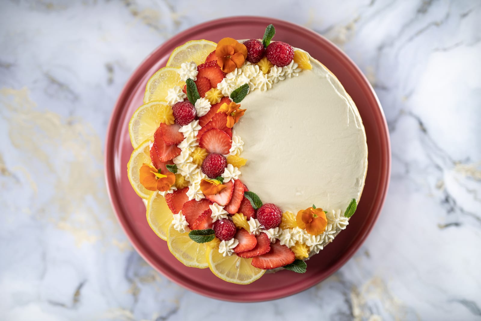 A creamy lemon cheesecake topped with lemon slices, berries, cream and orange flowers on a dark pink plate, from above