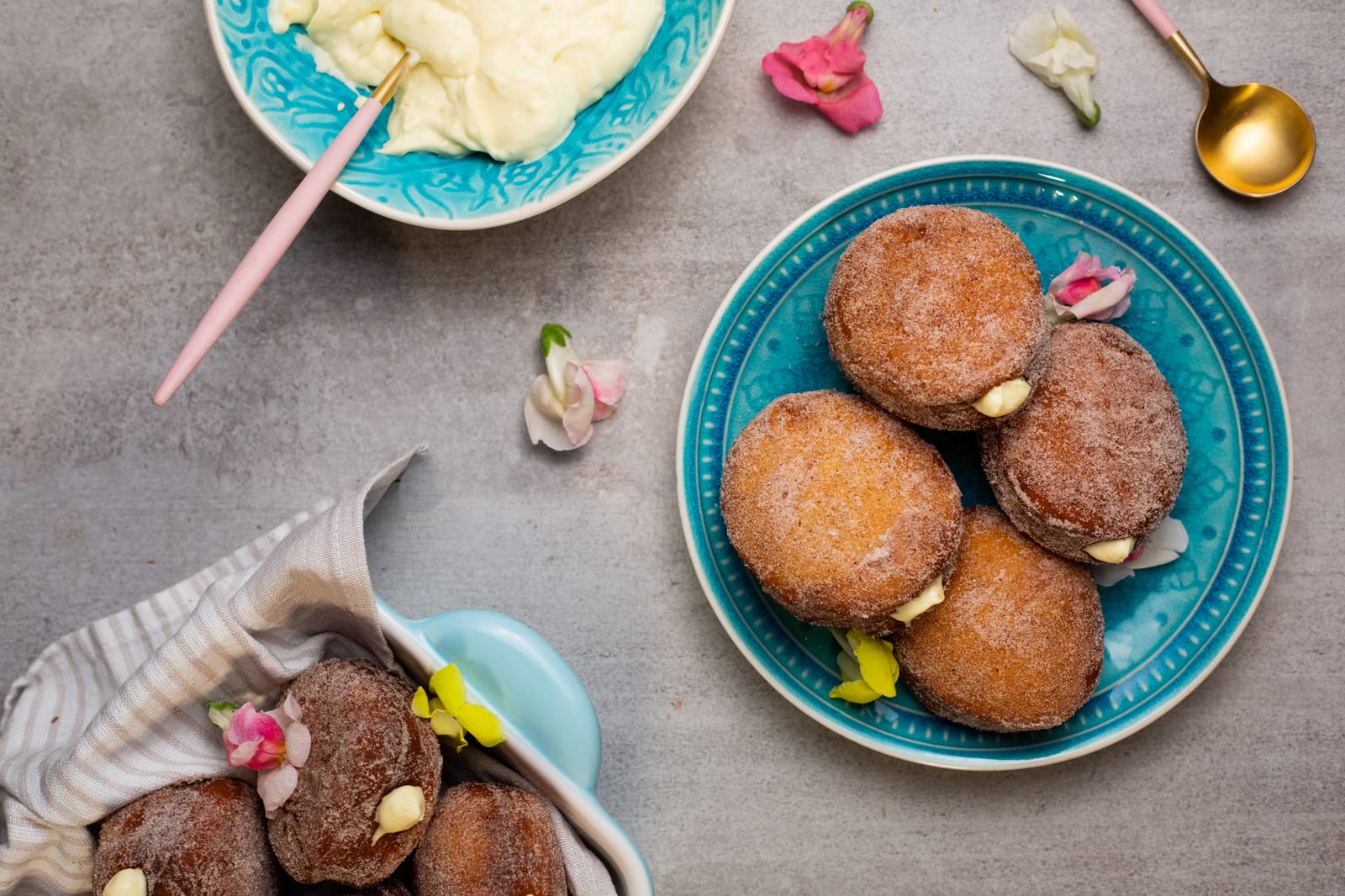 Sugared doughnuts filled with milk tart mousse on a teal plate beside a bowl of mousse and spoons, from above