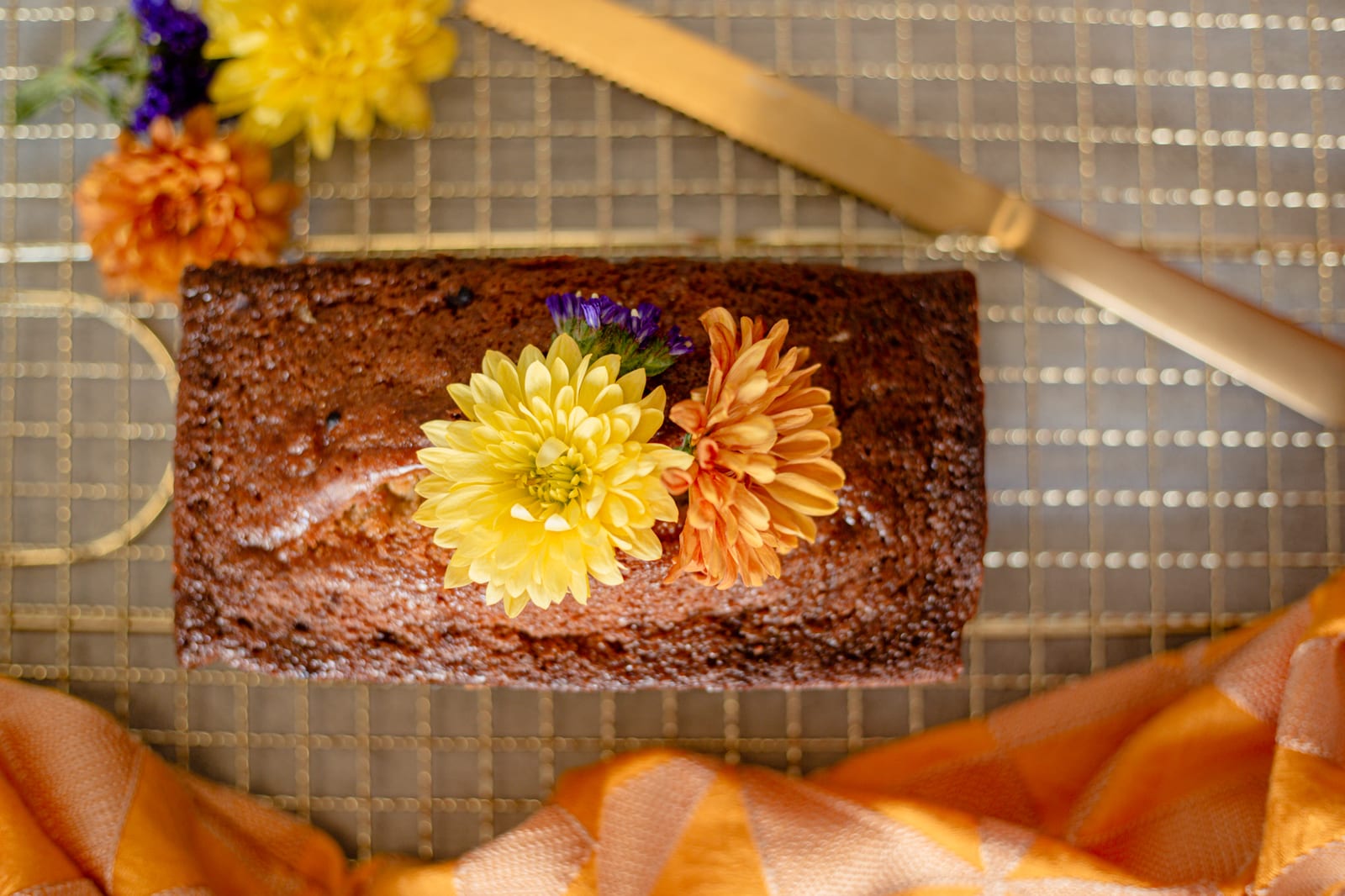 A lemon and blueberry loaf with yellow and orange chrysanthemums on a gold wire rack and orange silk, from above