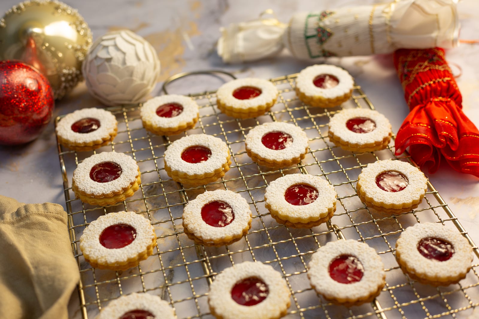 Rows of round lemon and raspberry Linzer cookies on a gold wire rack, with Christmas baubles and red ribbon