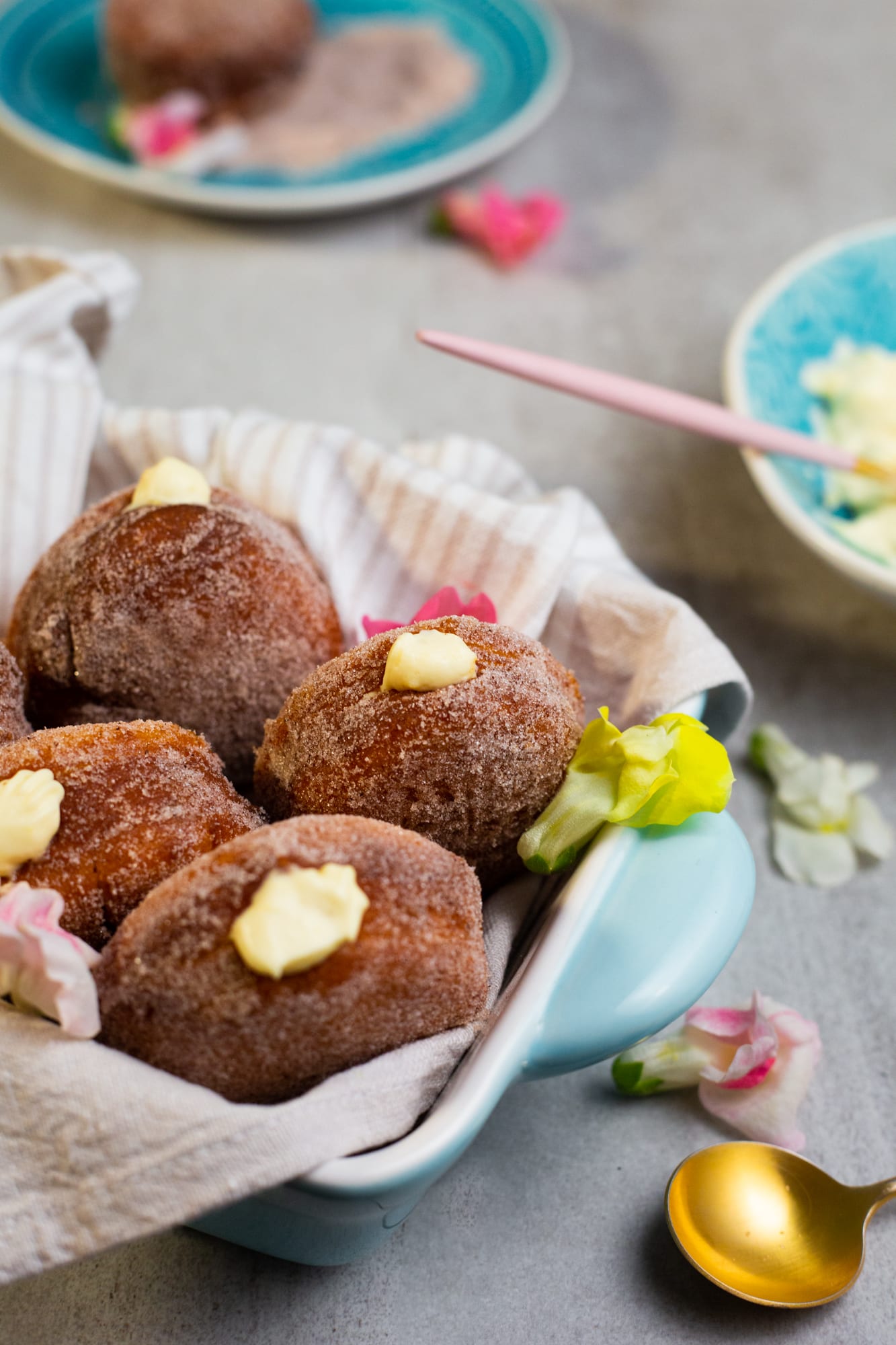 Cinnamon-sugared doughnuts filled with milk tart mousse in a cloth-lined dish, with a gold spoon and petals