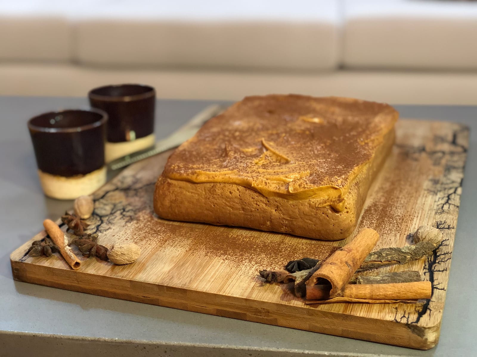 A masala tres leches cake dusted with spice on a patterned board beside cups and cinnamon sticks