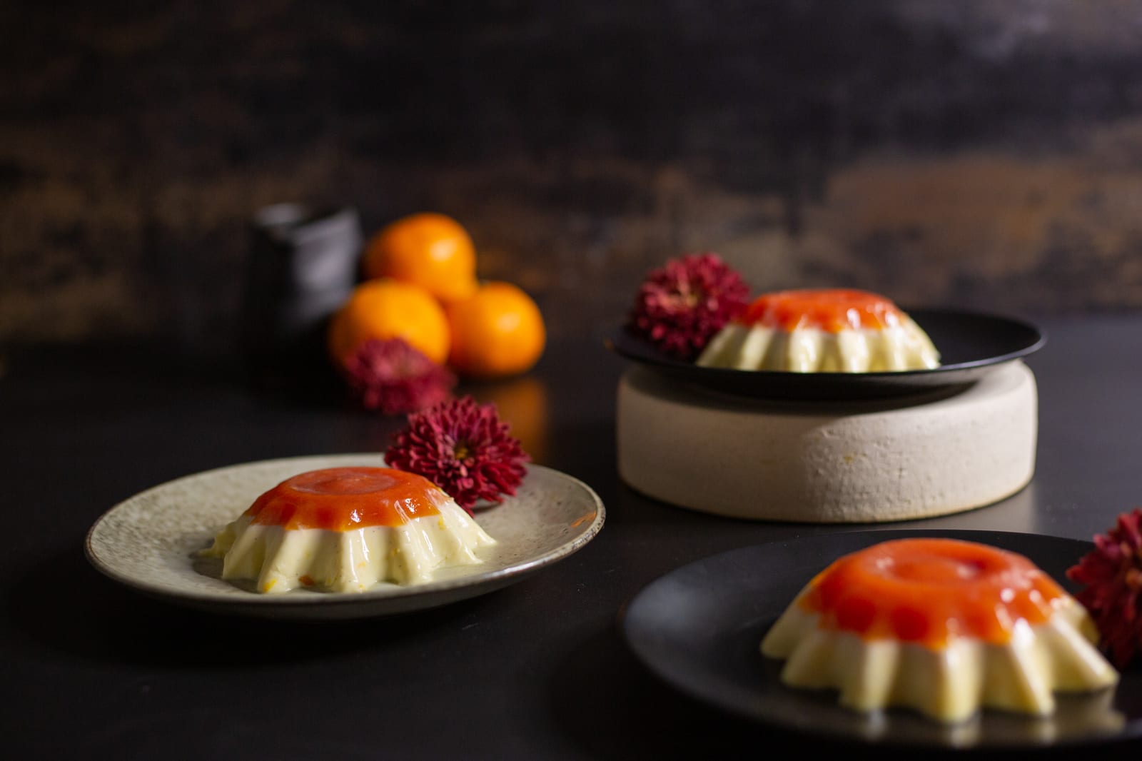 Three vanilla yoghurt panna cottas with blood orange jelly on dark plates, with oranges and red chrysanthemums