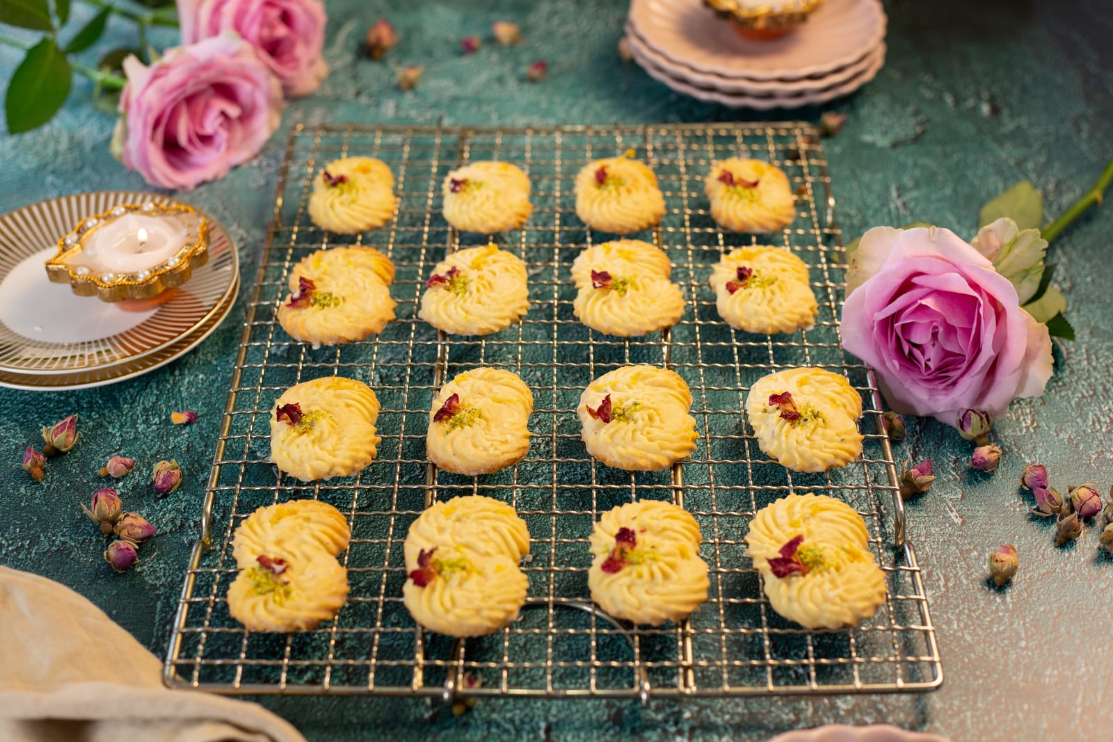 Rows of piped lemon, rose and cardamom biscuits on a wire rack, with pink roses and diyas