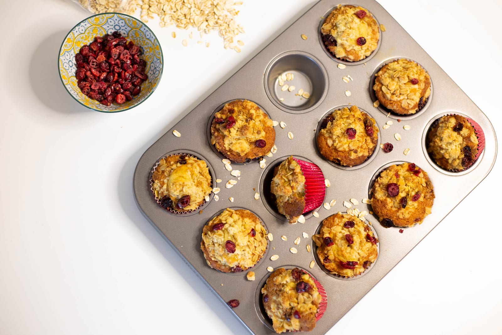 A tin of cranberry crumble muffins with a bowl of dried cranberries and scattered oats, from above