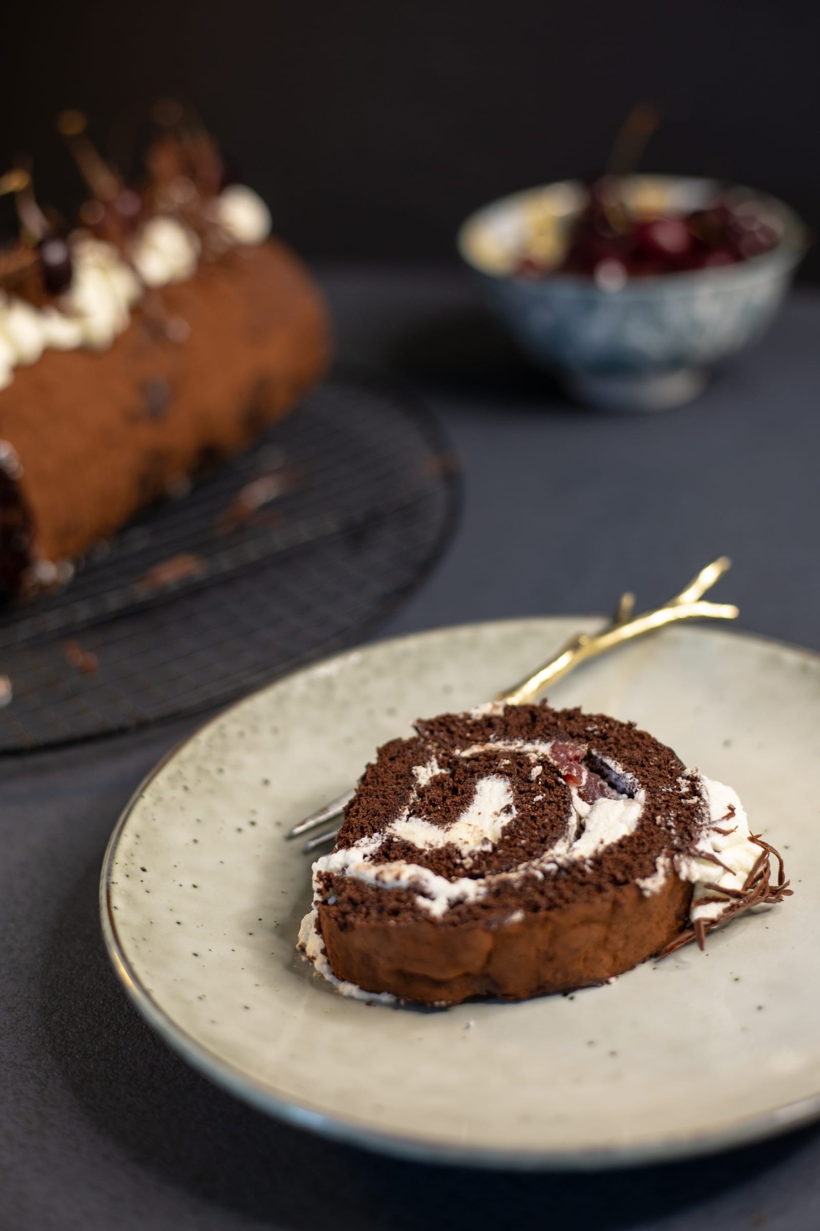 A slice of Black Forest Swiss roll on a cream plate with a fork, the roll behind