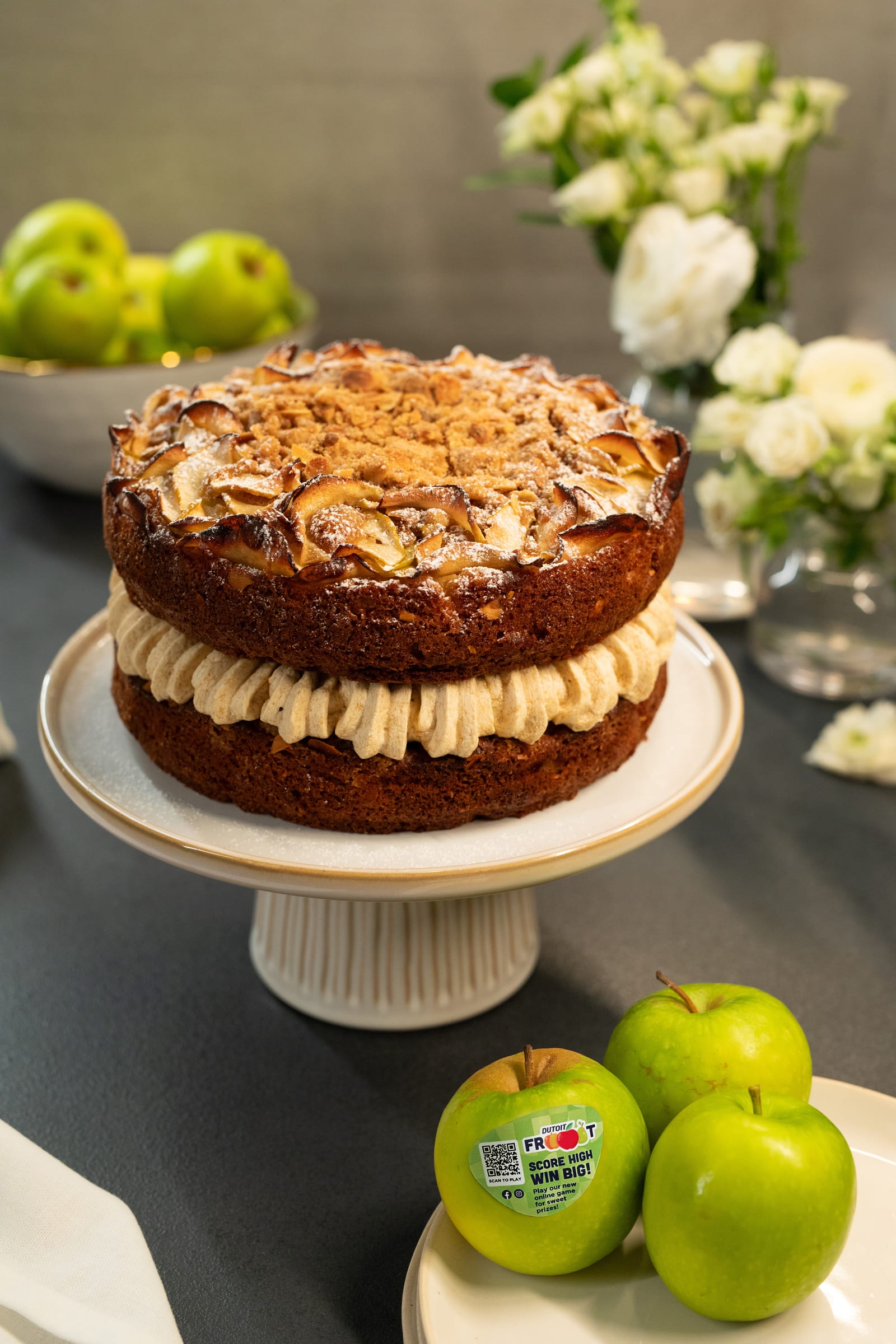 An apple crumble cake on a white cake stand with Granny Smith apples and white flowers