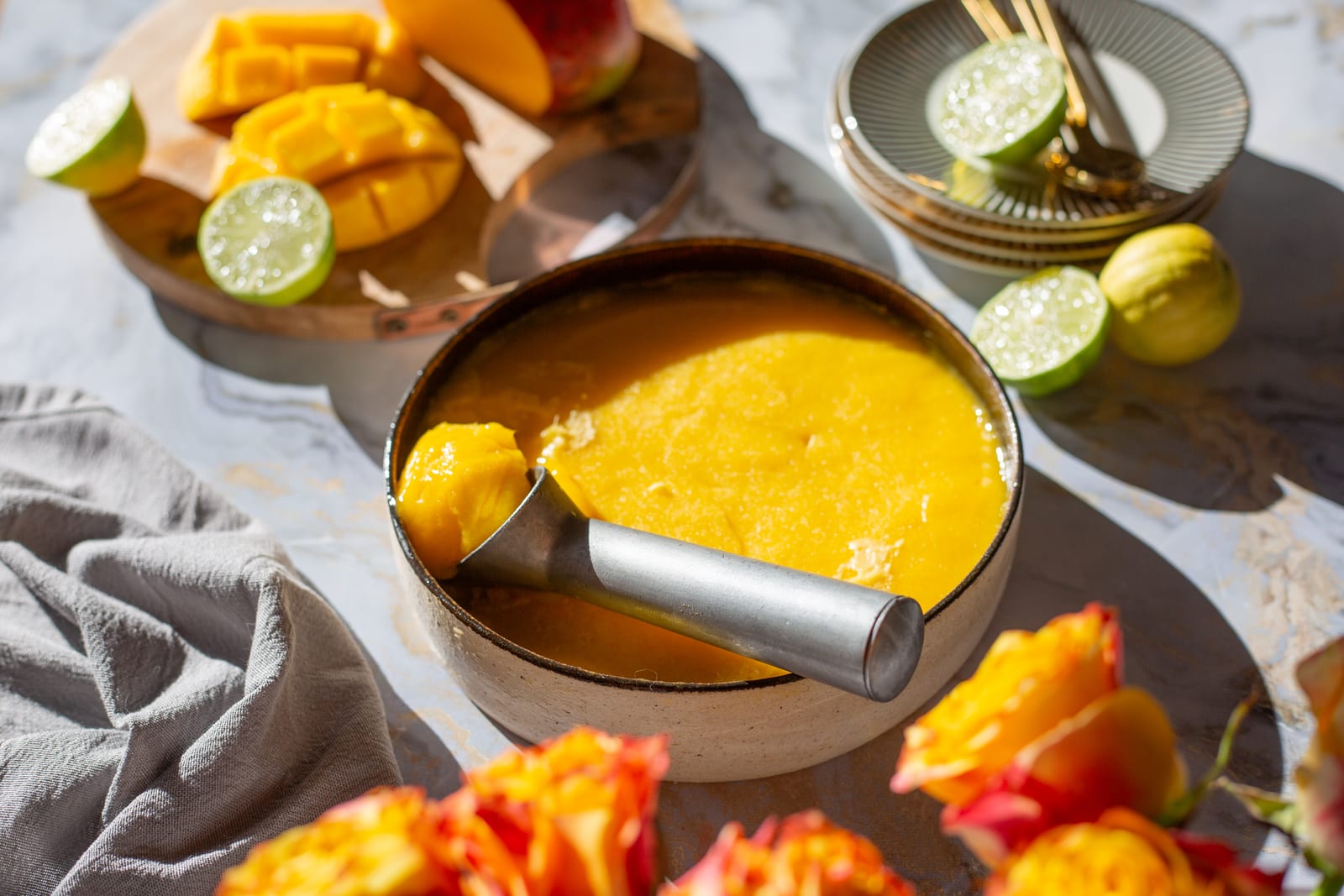 A dish of mango and chilli sorbet with an ice cream scoop, among cut mango, limes and orange roses
