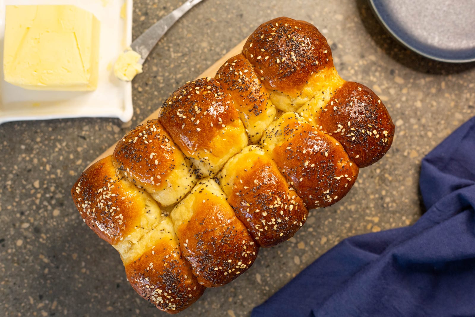 A glossy mosbolletjies loaf on a wooden board next to butter, from above