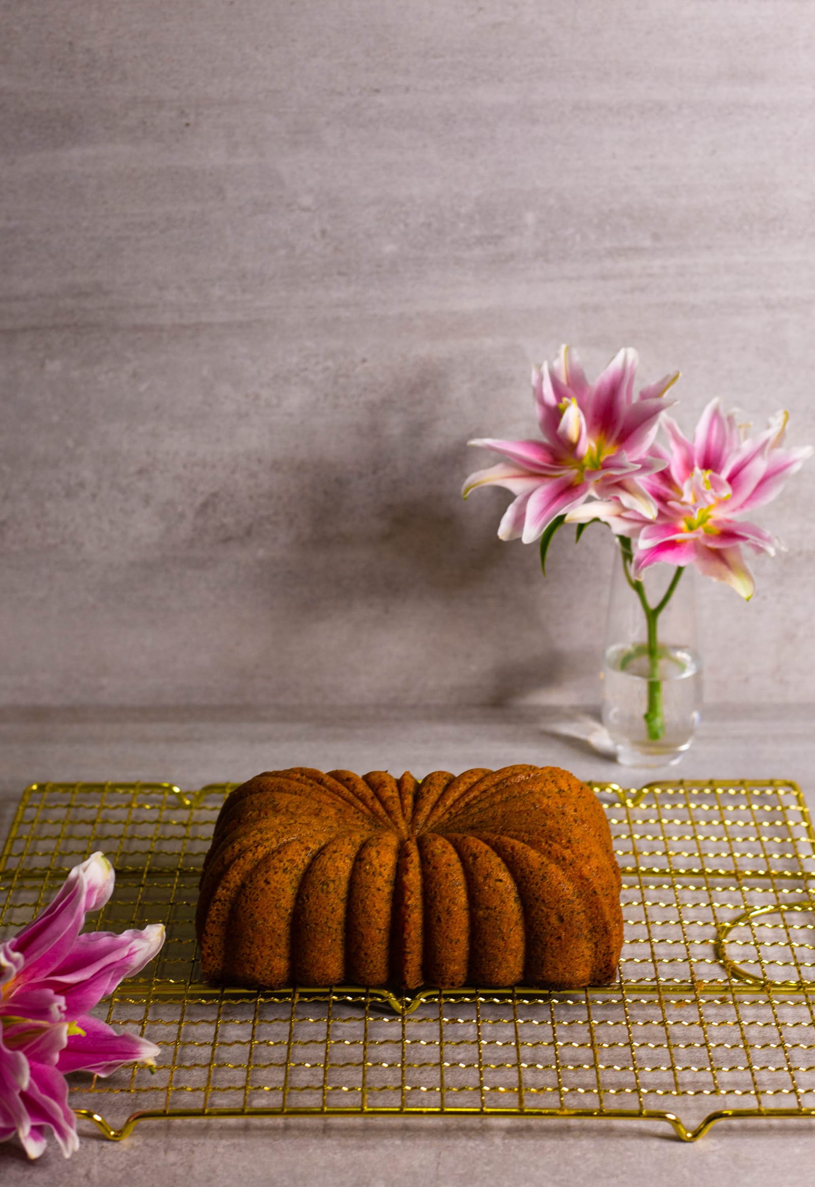 An unglazed gluten-free lemon and poppyseed loaf on a gold wire rack, lilies in a vase behind