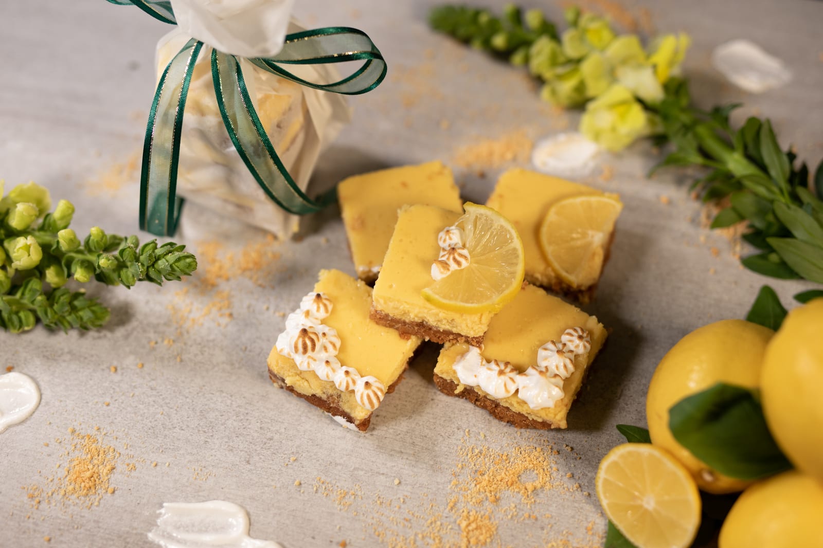 Sweet and tart lemon squares beside a gift bag tied with green ribbon, snapdragons and lemons