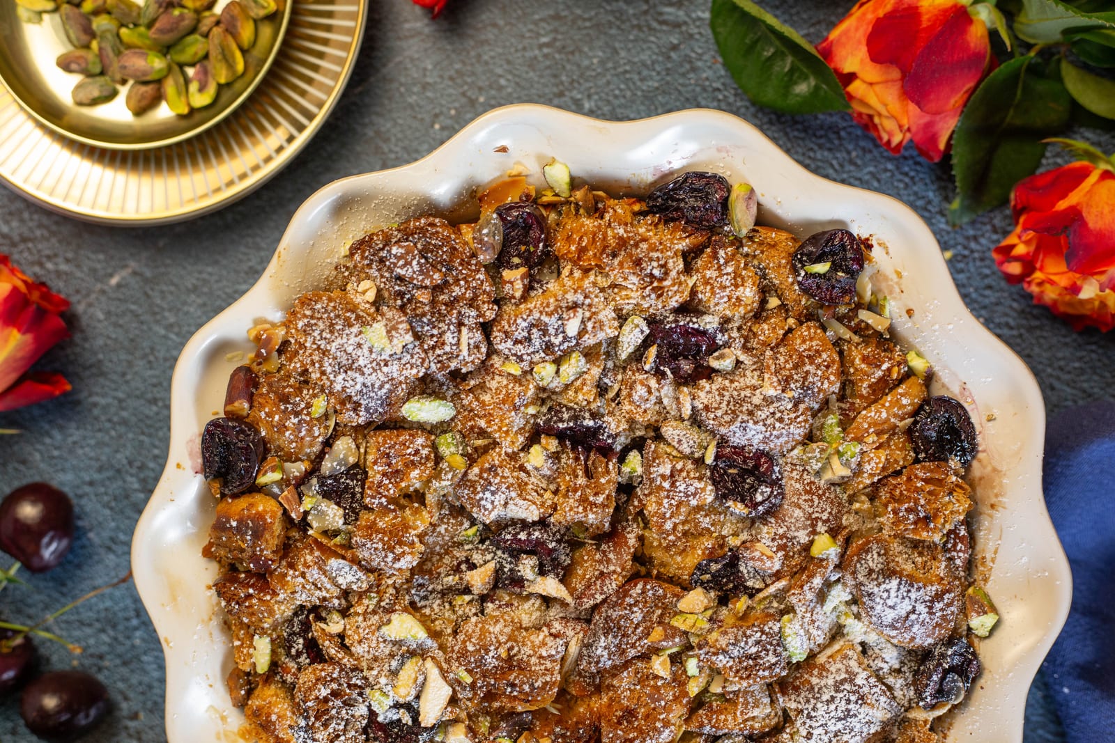 Chai-spiced bread and butter pudding from above in a scalloped white dish, scattered with pistachios, cherries and icing sugar