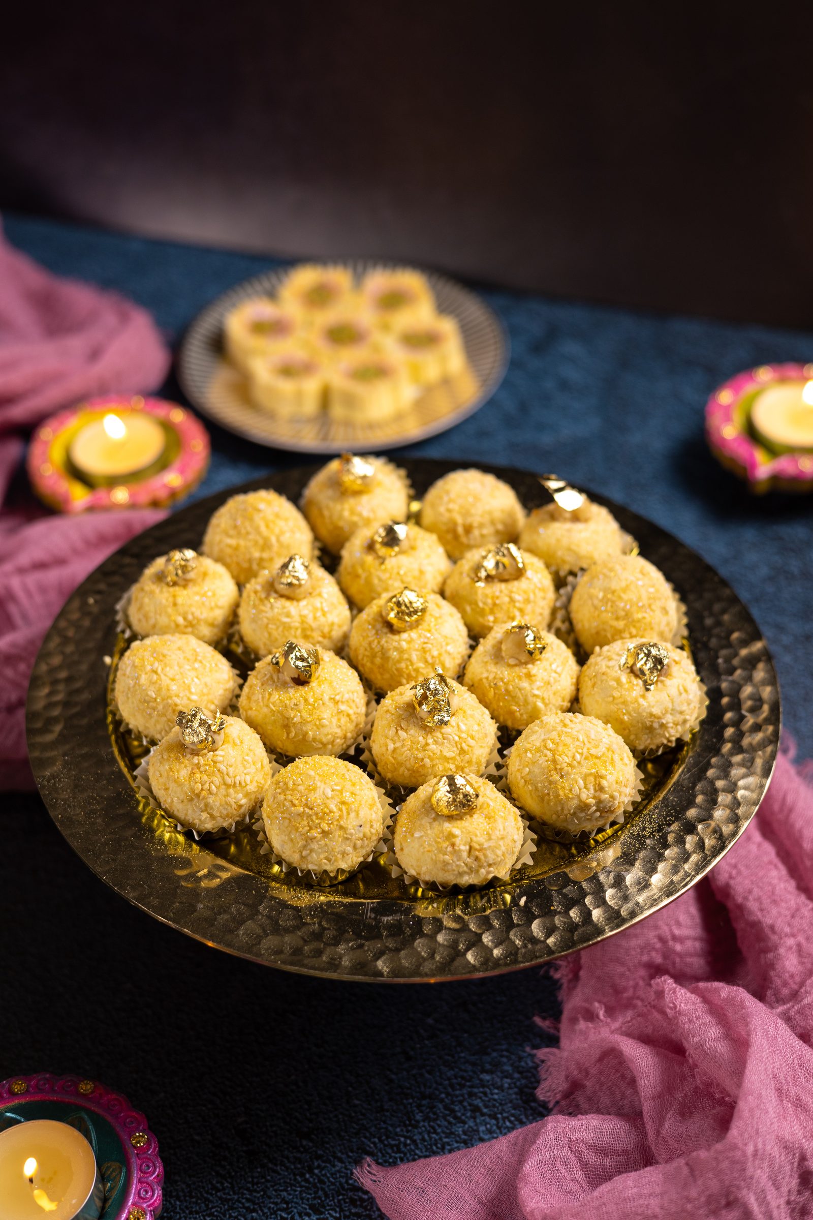 A hammered copper platter of coconut Simply Wonderfuls with a plate of flower-shaped Simply Wonderfuls and diyas behind, from the side