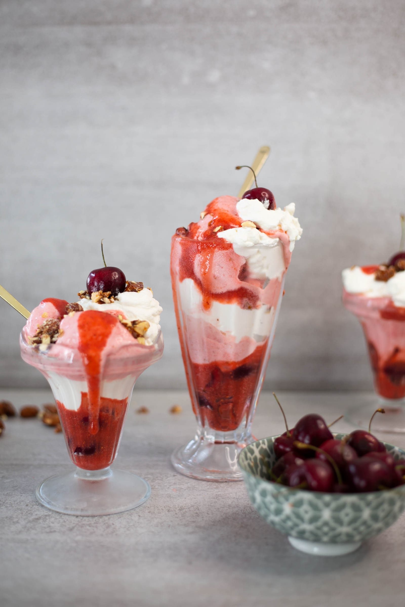 Three berries and cream sundaes of different heights beside a bowl of cherries