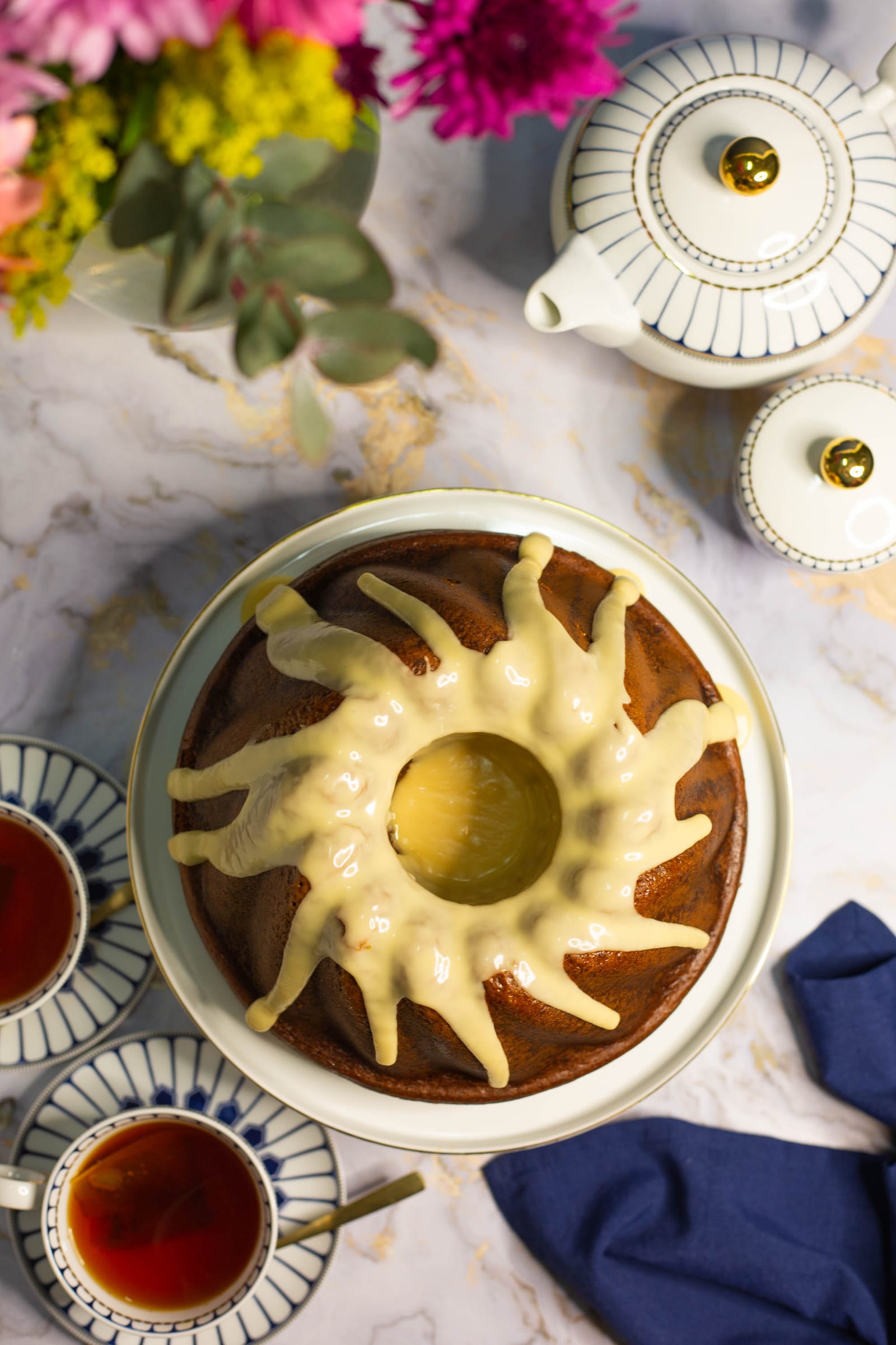 A rooibos and honey bundt with a sunburst of white glaze beside a cup of tea, from above