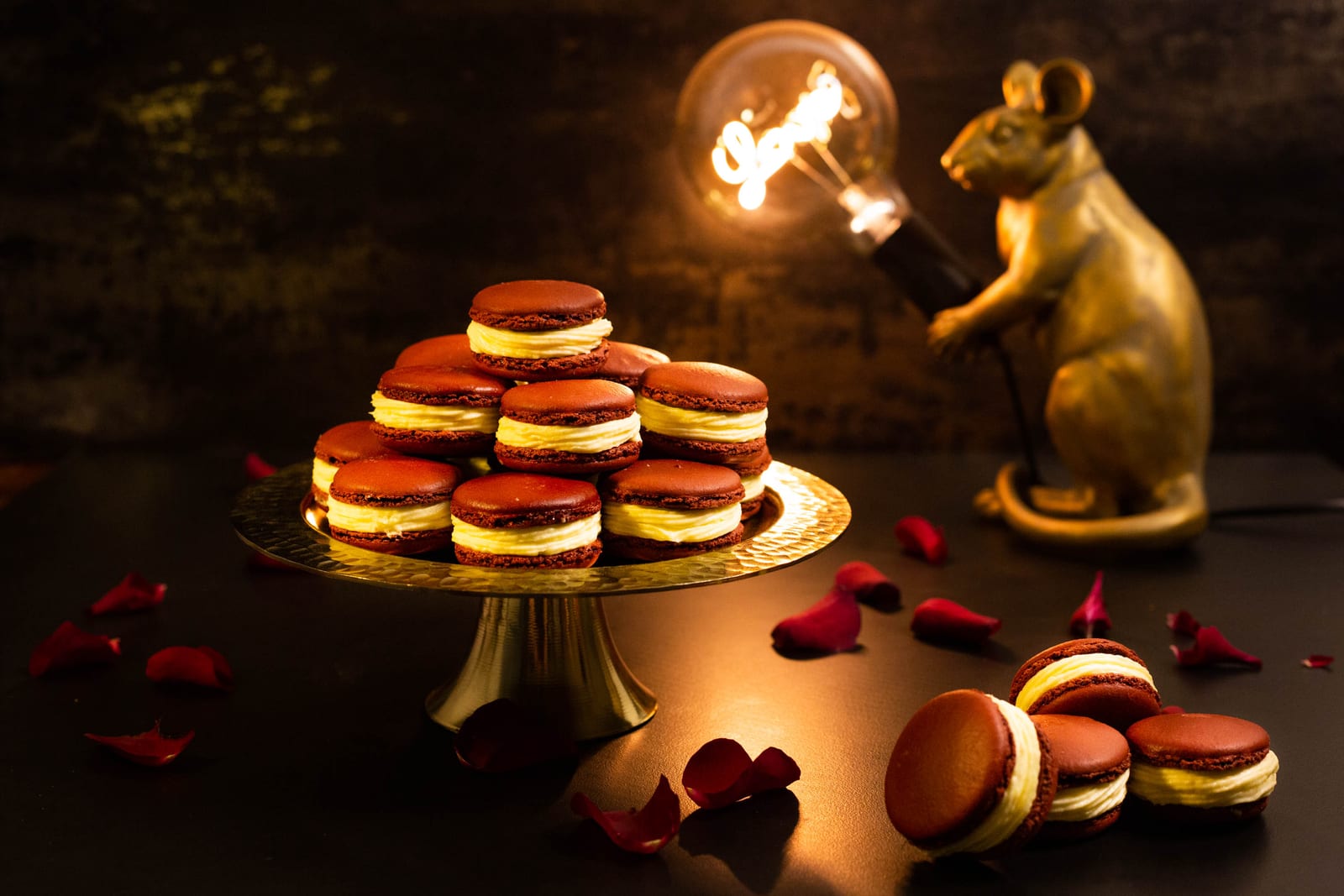 Red velvet macarons stacked on a gold stand beside a gold lamp and rose petals