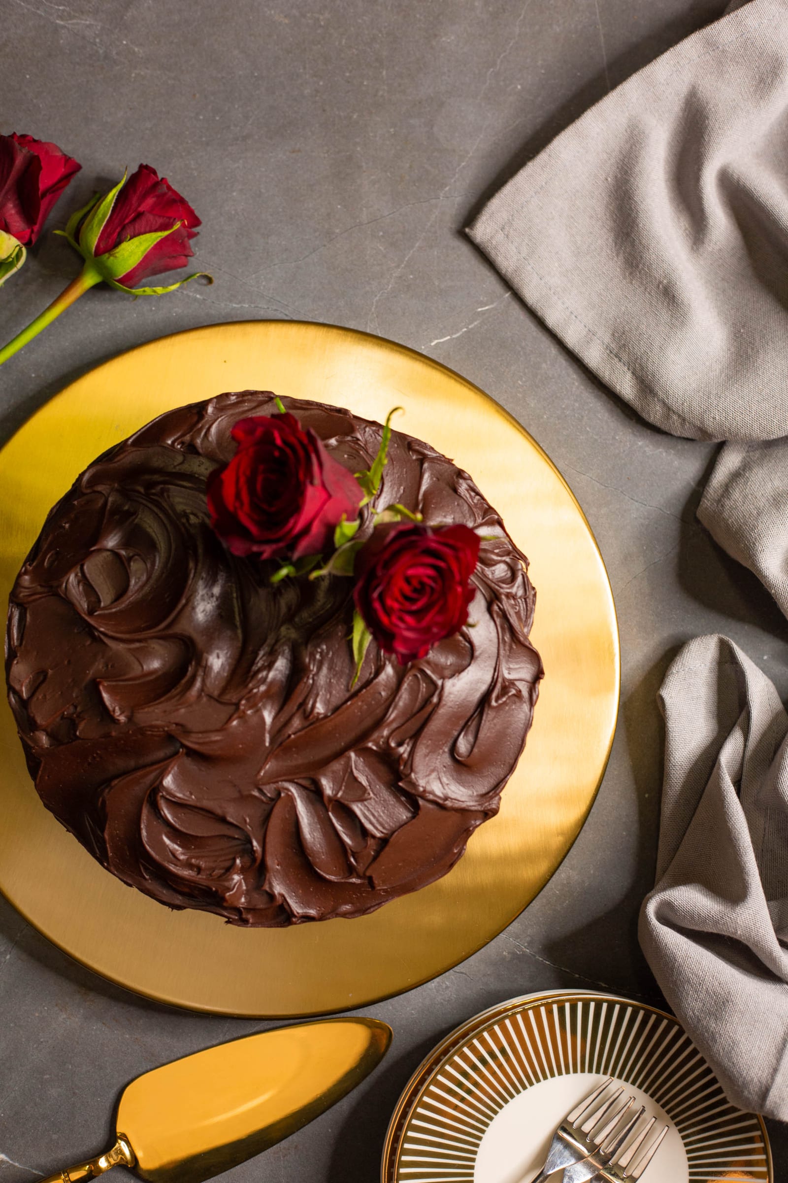 A chocolate and beetroot cake with glossy ganache and red roses on a gold board, plates and forks beside it, from above