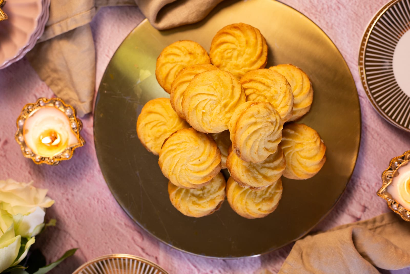 Orange and almond melting moments piled on a gold plate with lit diyas and white roses on pink linen, from above