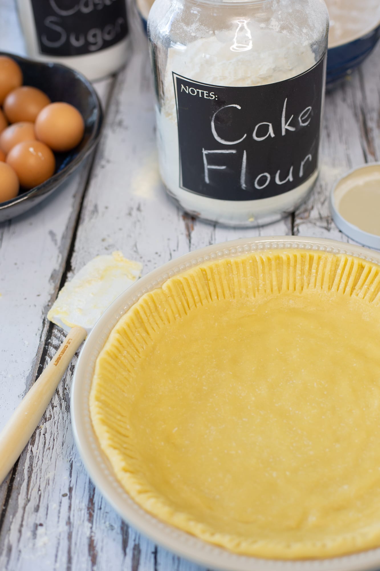 The unbaked pastry case for melktert in a pie dish beside a jar of cake flour and eggs