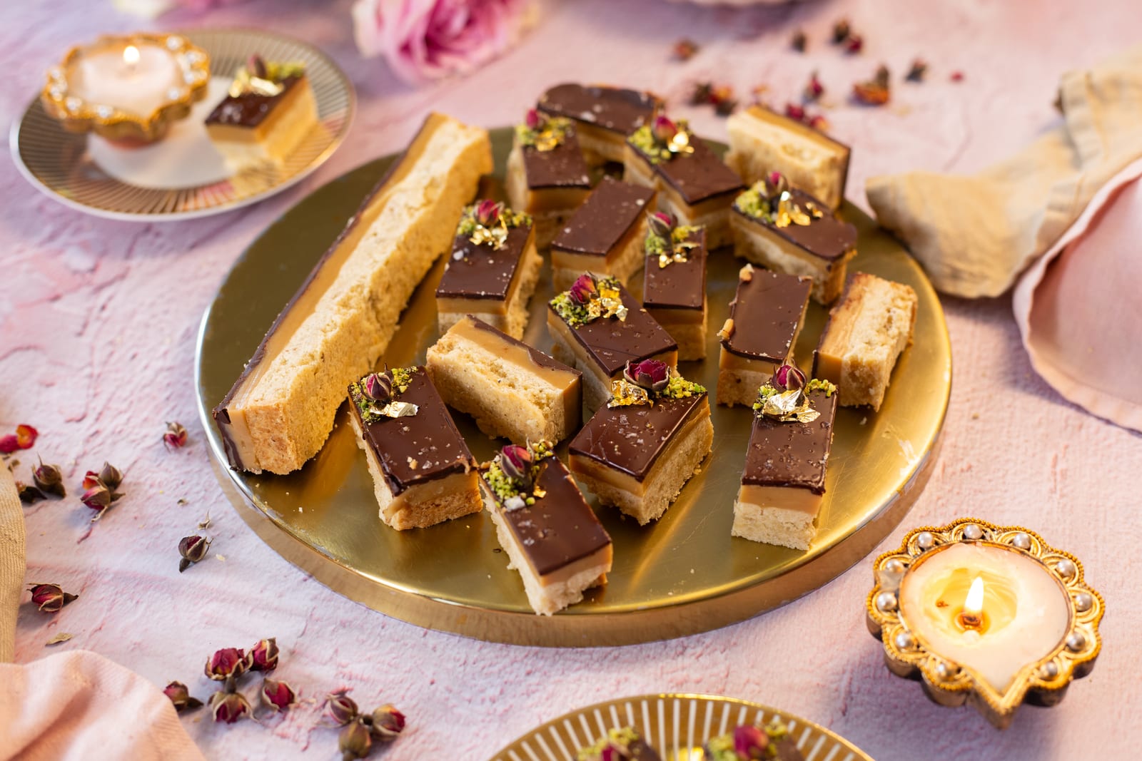 Squares of chai-spiced millionaire's shortbread on a gold platter, with a lit diya and rose buds