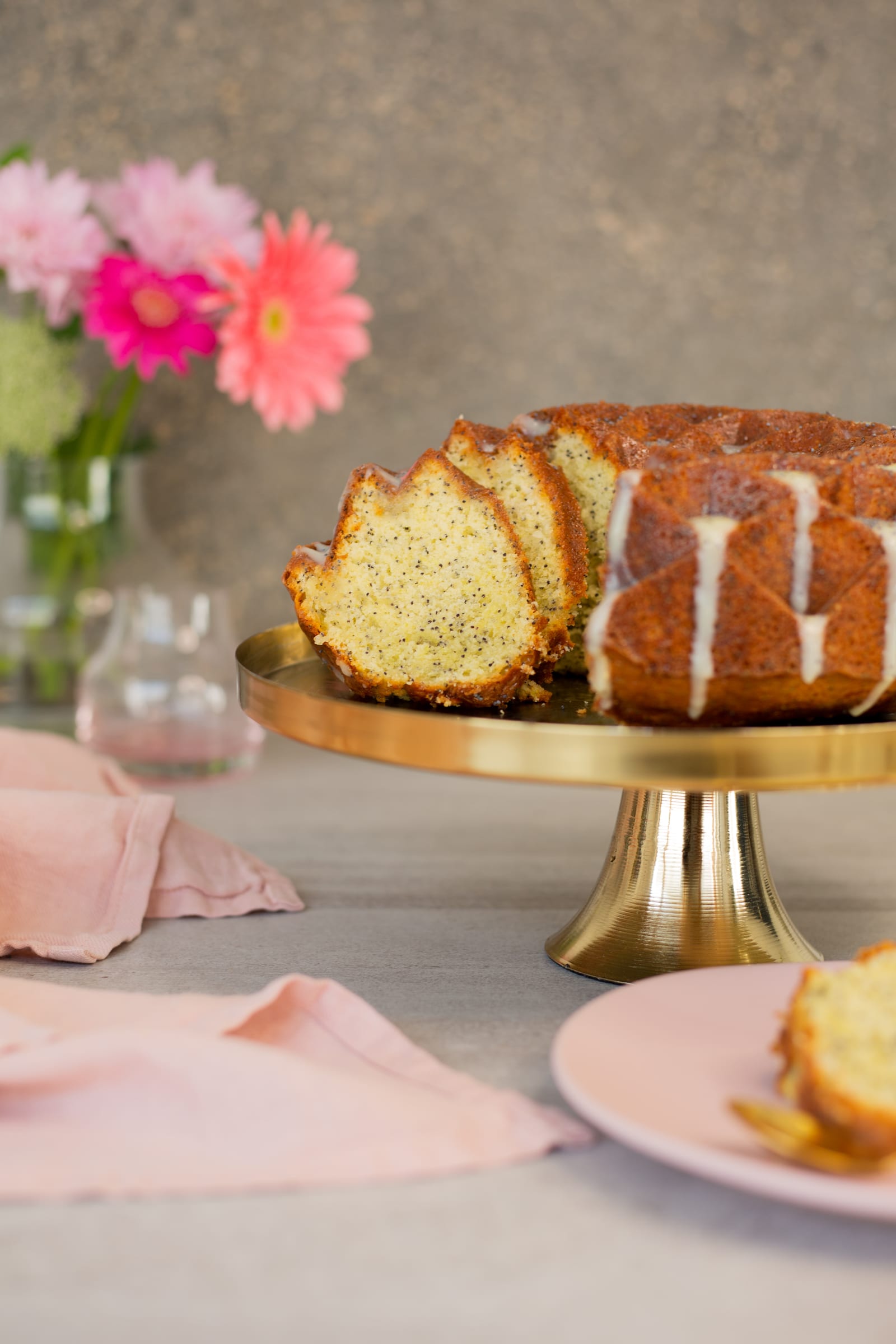 The cut lemon, poppyseed and olive oil bundt on its stand, a slice on a pink plate in front