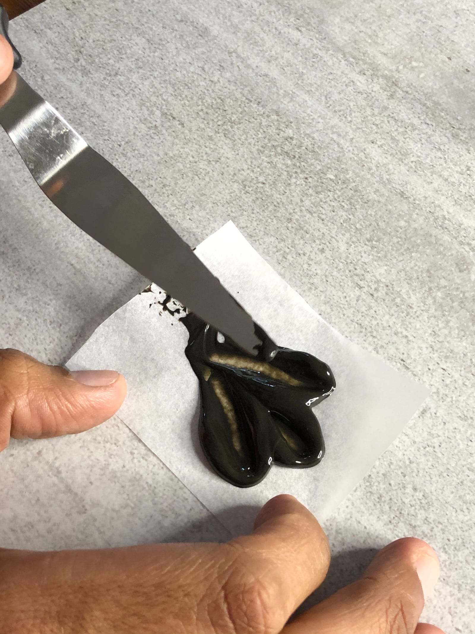 A three-lobed petal being spread in dark chocolate with a palette knife on a square of baking paper