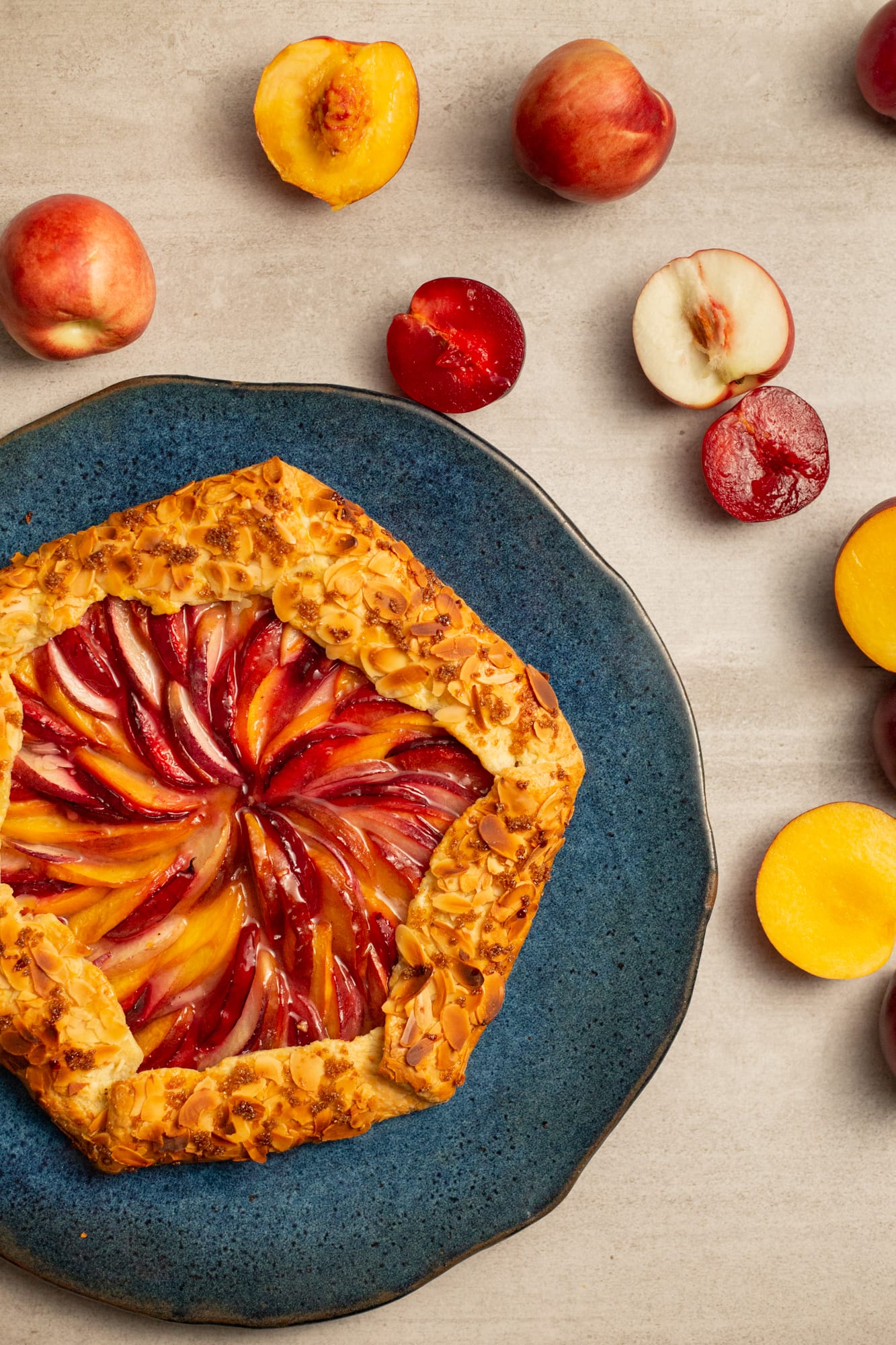 A whole baked stone fruit galette on a teal plate with halved plums and apricots, from above
