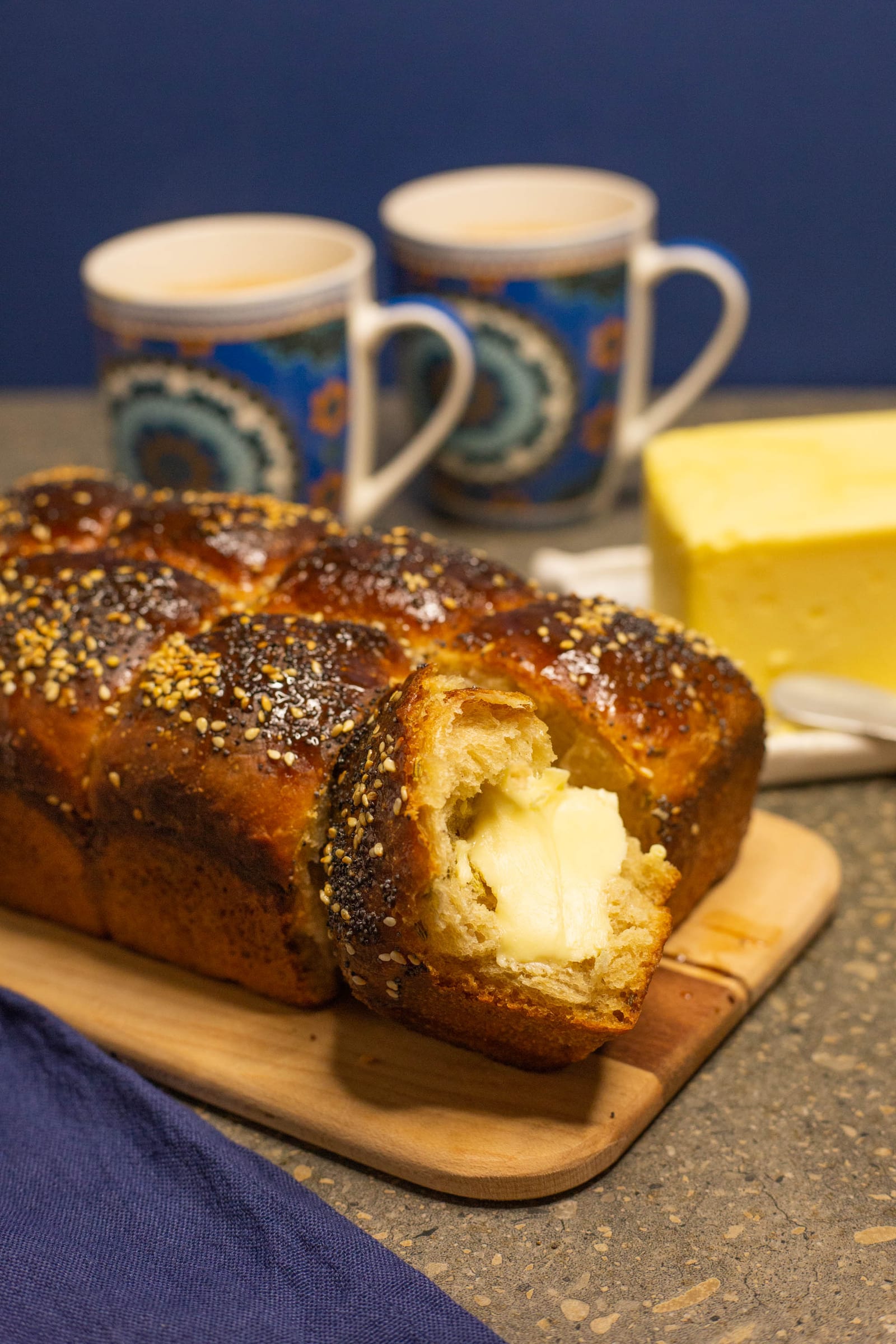 Seeded mosbolletjies with one end torn open and spread with butter, mugs of tea behind