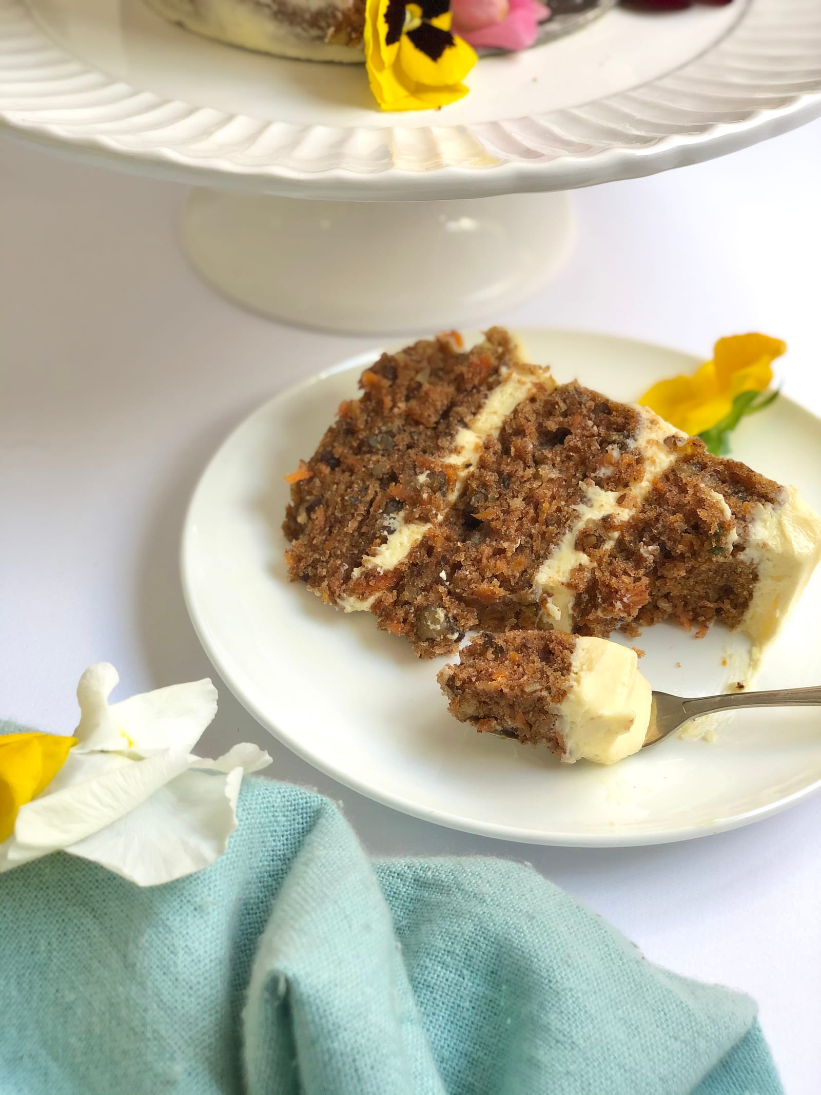 A slice of carrot cake on a plate with a fork under the cake stand, pansies around