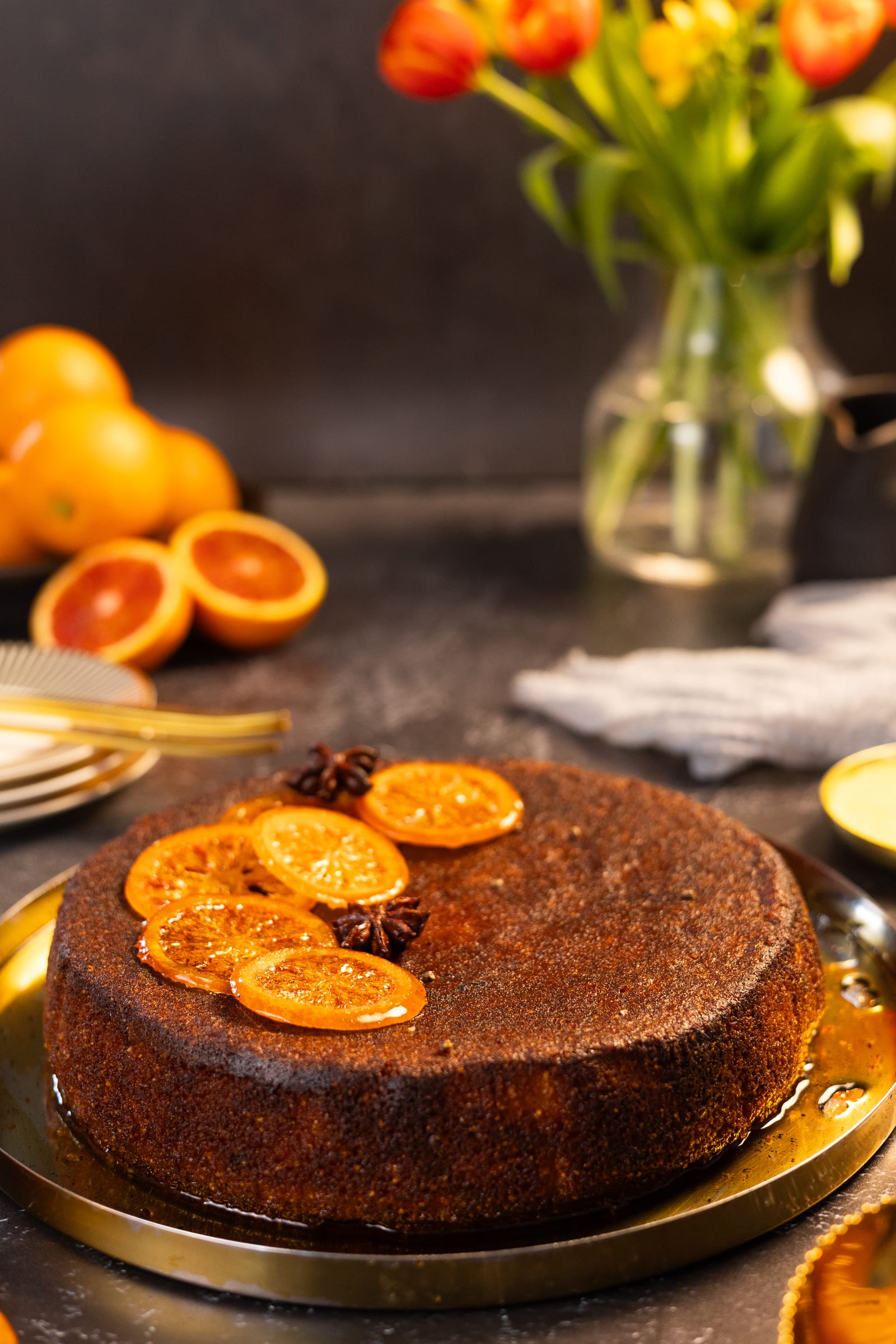 The blood orange polenta cake on a gold stand, with tulips and oranges behind